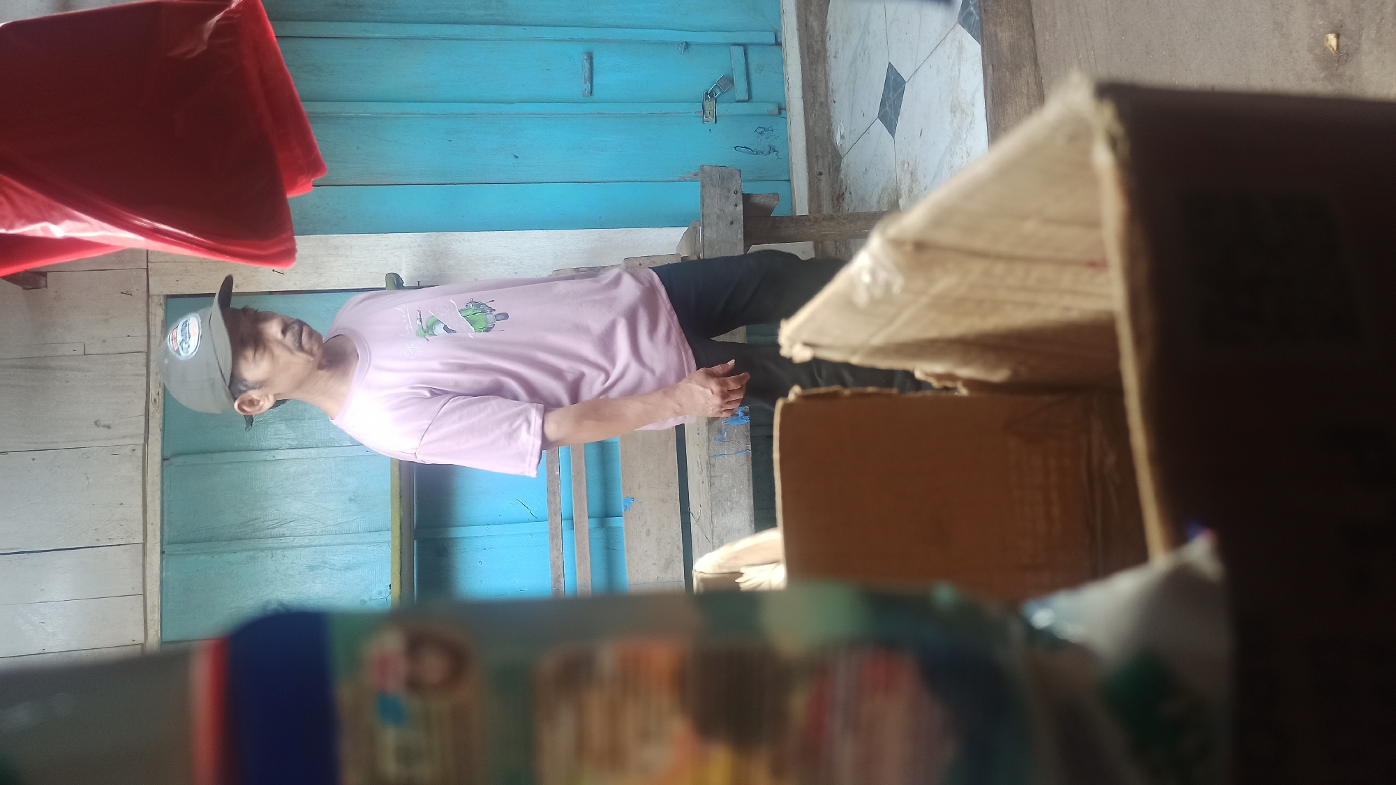 The image depicts a man standing in what appears to be an outdoor area with a wooden wall and a blue door in the background. He is wearing a pink t-shirt with a graphic print and a green baseball cap. In the foreground, there are cardboard boxes, one of which is in focus and partially obscuring a blurry object on the left. The scene suggests a casual, possibly commercial or residential, setting. The location is identified as Trisinar, Indonesia.