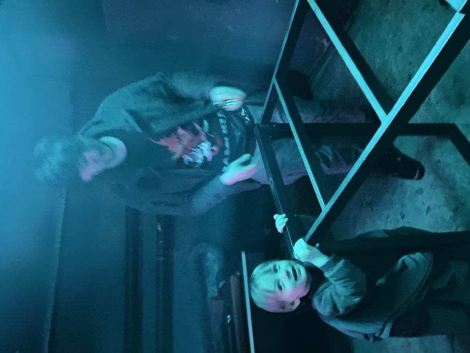 A man and child interact with a metal structure. The man stands wearing a dark shirt with a graphic and dark pants. The child, wearing a long-sleeved shirt and pants, grasps the structure. The setting is dimly lit with blue and green hues, creating a mysterious atmosphere. The metal structure features a grid-like pattern. The overall composition has a playful and curious tone, focusing on interaction within an intriguing environment.
