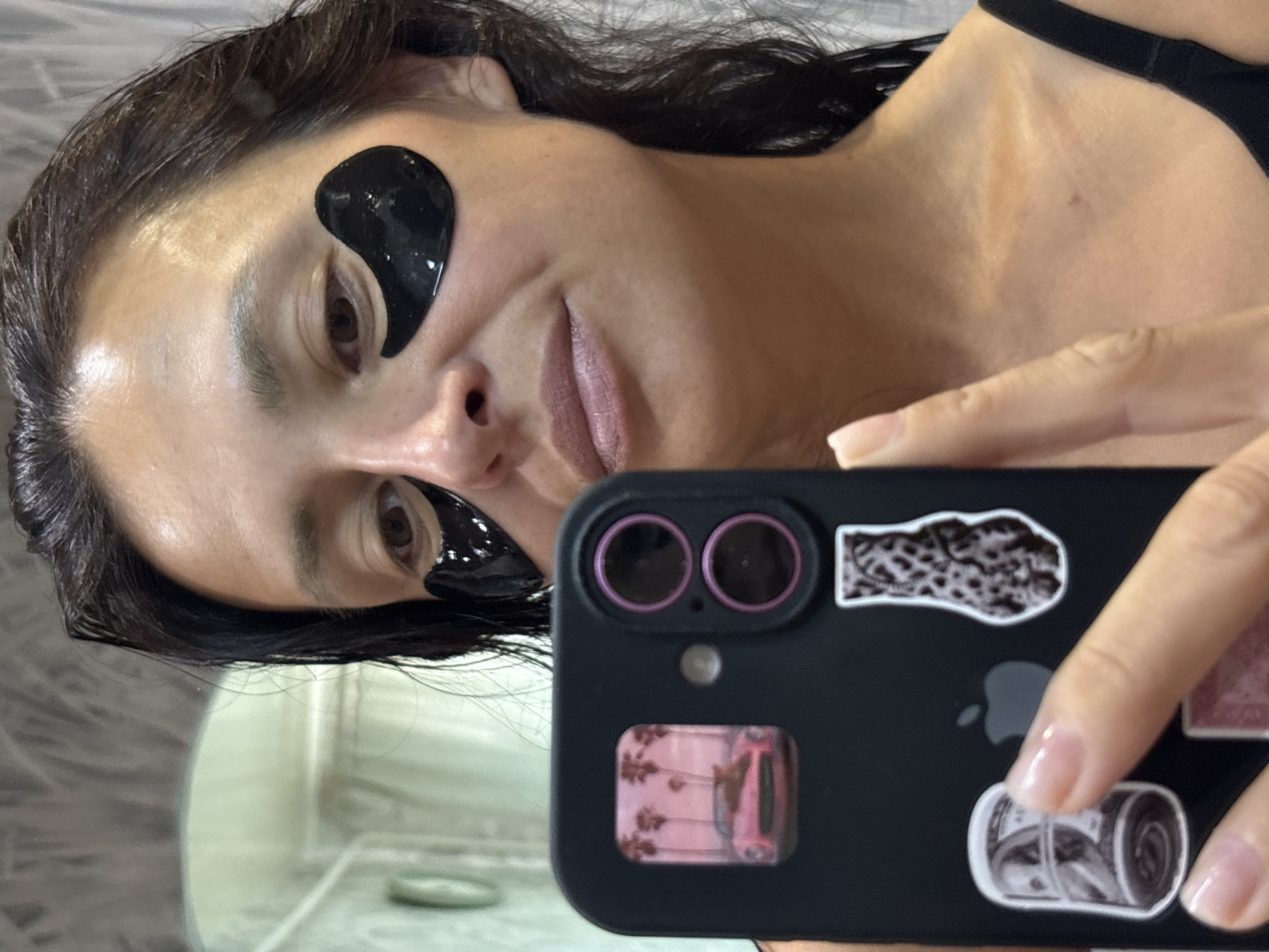 The media file displays an image rotated approximately 90 degrees counter-clockwise, capturing a close-up selfie of a woman engaged in a skincare routine. She is seen from the chest up, with her dark hair framing her face. She has two glossy black under-eye patches applied below her eyes. Her expression is a soft, subtle smile, and she looks towards the camera. Her left hand holds a black smartphone, featuring a dual-lens camera setup with distinct pink or purple rings around the lenses. The phone case is personalized with several stickers: a prominent Apple logo, a vibrant pink image depicting a vintage car beneath palm trees, a black and white sticker resembling a roll of film, and another abstract black and white textured design. She wears a black top or strap. The background is softly blurred, suggesting an indoor setting with light-colored walls or surfaces, possibly a bathroom or bedroom. No direct geographical cues are discernible in the image, but the provided context places the image in Chaikovsky, Russia. The overall impression is one of casual self-care.