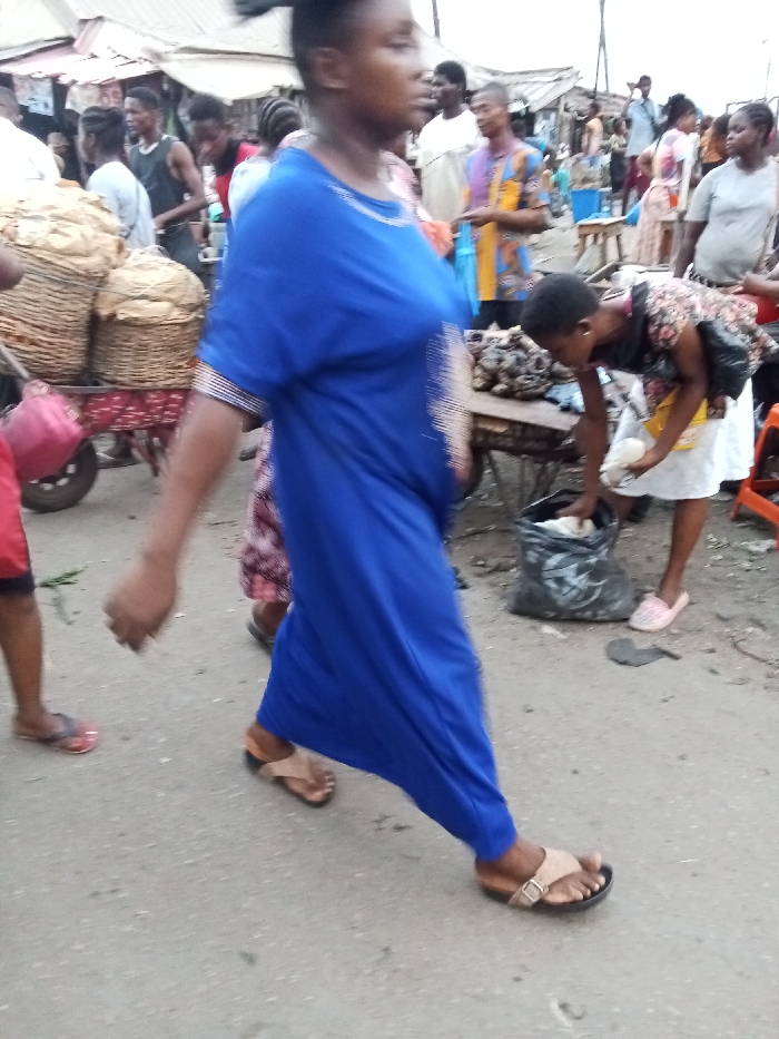 The image portrays an outdoor market scene on a paved surface in Owerri, Nigeria. In the immediate foreground, a dark-skinned individual, likely female, wearing a royal blue long dress with light embroidery and tan buckled sandals, walks towards the right. Her head is turned slightly to the right. To the right of this central figure, a young dark-skinned female, dressed in a floral top, white skirt, and pink flat shoes, bends forward. She is reaching into a black plastic bag positioned on the ground with her right hand, while holding a small white object in her left hand. A makeshift wooden table directly in front of her displays numerous dark, irregularly shaped items.
In the far left foreground, the lower torso and legs of a person wearing red shorts and red flip-flops are visible, holding green leafy material. Further into the mid-ground on the left, a pushcart laden with several large woven baskets, which contain bundled goods, is visible. The background is populated by numerous dark-skinned individuals, including a male wearing a multi-colored patterned shirt, standing amidst various market stalls topped with corrugated metal roofs. The scene conveys a bustling commercial environment.