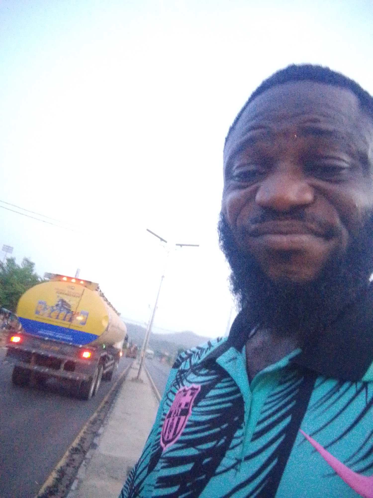 A man with dark skin and a full beard is taking a selfie in what appears to be Lokoja, Nigeria. He's wearing a turquoise and black patterned shirt with a pink Nike logo and a crest that resembles the FC Barcelona logo. Behind him, a yellow tanker truck with blue lettering is driving down a road bordered by a curb. Street lights are visible in the background, and the sky is overcast.