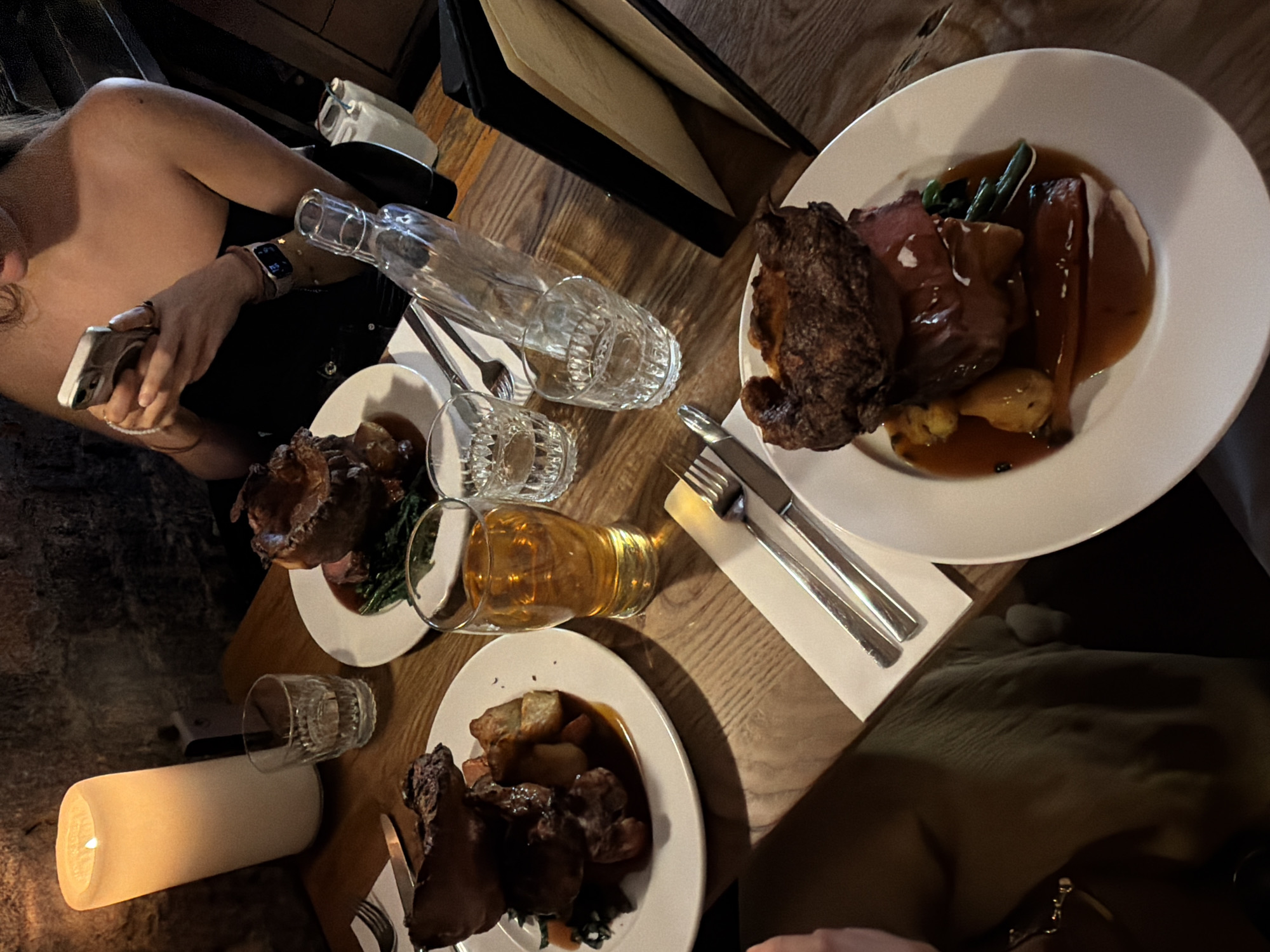 This image depicts a dinner table scene, seemingly indoors, at an unknown location in an unknown city. Two individuals are partially visible, one on the left using a smartphone and the other on the right. The table is set with three plates of food, potentially roast dinners, each containing meat, vegetables, and gravy. Additionally, there are glassware items including a water bottle, two smaller drinking glasses, and a glass of amber-colored liquid, likely beer or cider. Cutlery is neatly arranged beside the plates. The table is lit by a cylindrical candle on the left. In the background, a menu is visible. The overall lighting is dim, suggesting an intimate dining setting.