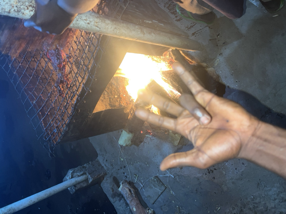 The image displays a nighttime outdoor scene in Birnin Kebbi, Nigeria, centered on a rudimentary barbecue setup. A metal grill stand, possibly fabricated from a repurposed barrel or similar material, is situated in the mid-ground. Within this stand, an active wood fire burns, emitting bright orange and yellow flames and illuminating the immediate area. Meat is visible cooking on a coarse wire grate positioned atop the stand, directly over the flames.
In the lower-left foreground, a dark-skinned human left hand is raised with fingers splayed, revealing a small dark mark on its palm. To the right of the grill, the lower leg and a foot wearing a green flip-flop are partially visible. A light-colored vertical pole is present on the right, held by a dark-skinned hand near the grill structure. Another similar light-colored pole extends upwards from a concrete base on the far left.
The ground surface is a mixture of packed earth and concrete, sparsely covered with small pieces of debris, including a bone-like object and a rectangular light-colored item. The background beyond the illuminated area is dark and indistinct.
