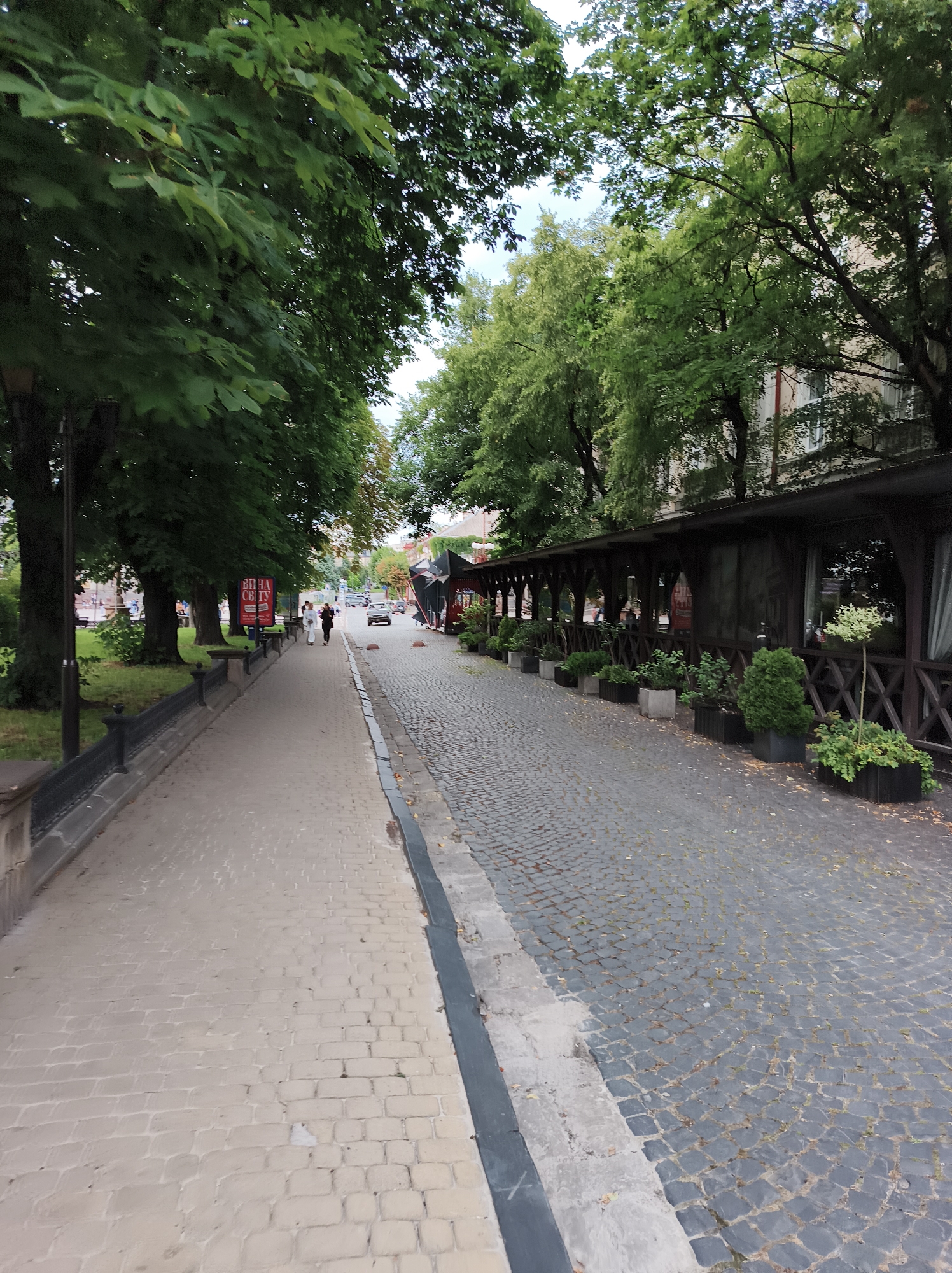 The image depicts a street scene in Ternopil, Ukraine, likely near a commercial area given the presence of an outdoor seating area for a restaurant or cafe. The foreground is divided into a paved pedestrian walkway on the left, constructed of light-colored brick pavers, and a cobblestone street on the right. The walkway is bordered by a black metal fence and a grassy area with large trees. On the right side of the street, there is an elongated, covered outdoor seating area with wooden supports and lattice work, featuring planters with greenery. Further down the street, a light-colored car is visible, and two individuals are walking on the pedestrian path. In the background, buildings and more trees can be seen, suggesting an urban environment. The overall lighting suggests it is daytime, with overcast or diffused light.
