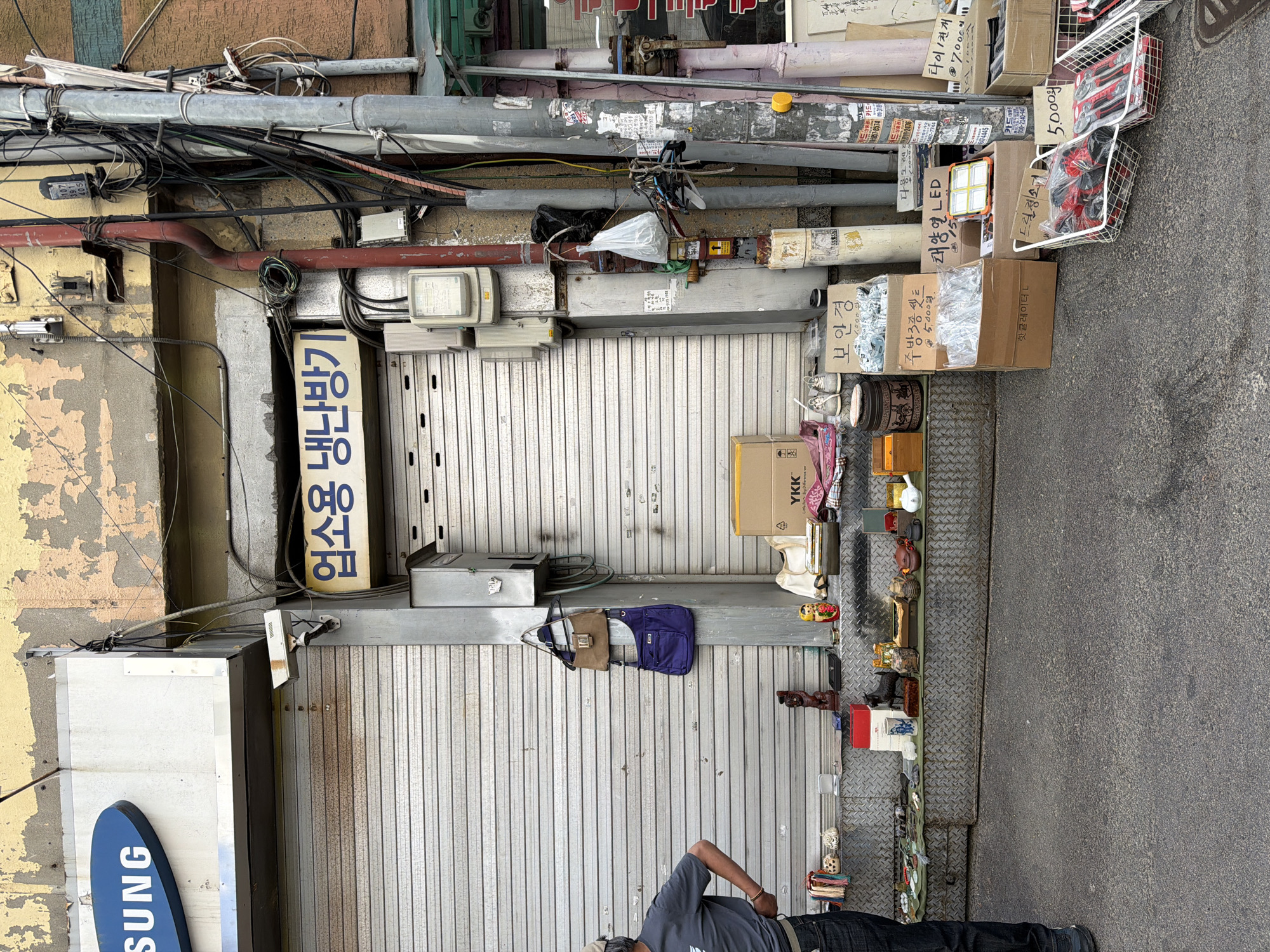 The image depicts a narrow alleyway in what appears to be Seoul, South Korea, with a closed metal storefront shutter. Various pipes and electrical wiring are visible along the wall above the shutter. Boxes and baskets containing goods, possibly for sale, are stacked along the edge of the shutter and extend onto the paved ground. A man is standing in the alleyway, looking at the items on display. The storefront has a sign with Korean writing and a partial "Samsung" sign.