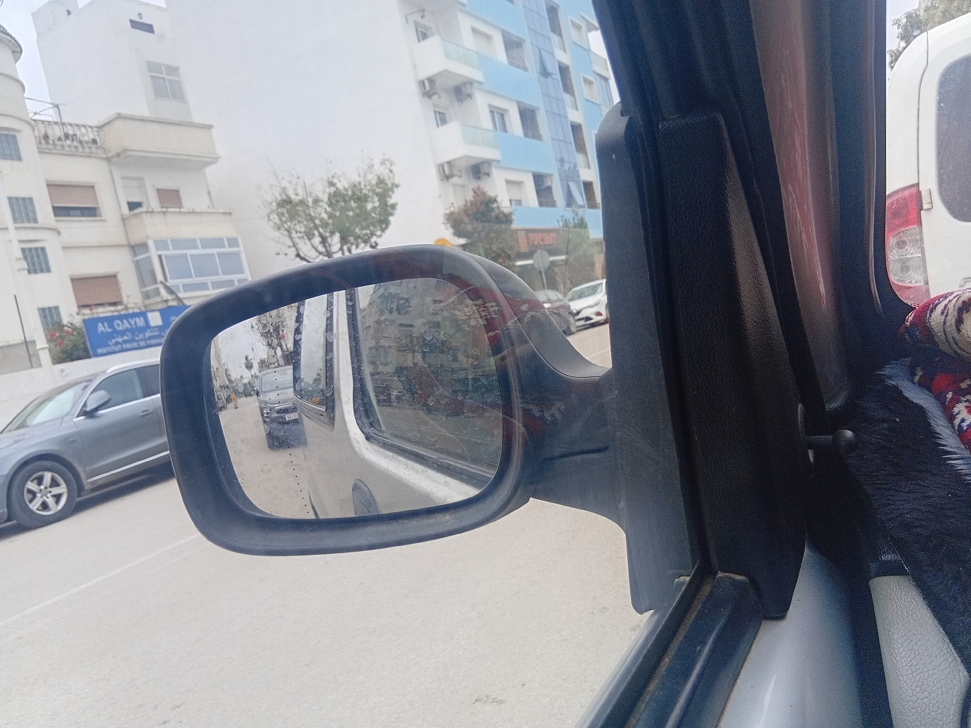 The image provides a street-level view from inside a vehicle, looking out the driver's side window and into the side mirror. The foreground features the car's black plastic door frame and a prominent side mirror.
Through the window on the left, an urban landscape unfolds under an overcast, hazy sky. Several multi-story buildings are visible, including a large white structure with balconies and a turret-like section on the far left. Below this building, a blue sign clearly displays "AL QAYM" in Latin script and "التكوين المهني" (Vocational Training) in Arabic, with "INSTITUT PRIVE DE FORMA..." partially visible, indicating a private vocational training institute. Further down the street, another building with alternating white and light blue panels is seen. Sparse, bare trees suggest a cooler season. A gray sedan, possibly an Audi, is positioned on the left side of the road, either parked or in motion.
The side mirror, framed in black plastic, reflects the scene behind the viewer's vehicle. In the reflection, a dark gray van or SUV is visible on the road, following the car. The mirror's surface appears somewhat streaked or dirty. To the far right, a portion of a white van's rear is visible outside the car window. Inside the vehicle, on the right, a colorful, patterned fabric, possibly a small rug or tapestry, is tucked into the window frame, adding a decorative element. The overall scene depicts a typical urban environment in Martil, Morocco, characterized by the architecture and visible text.