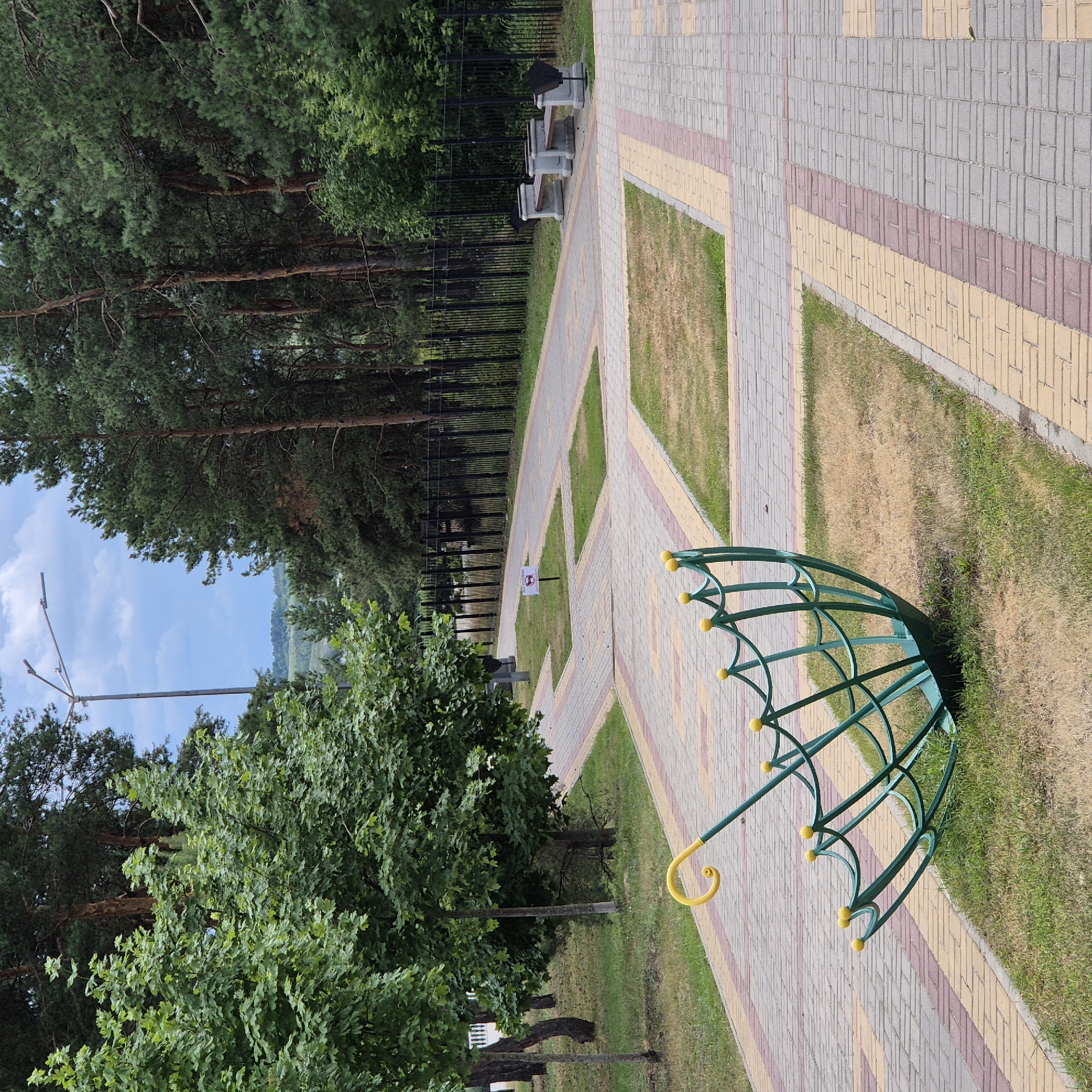 The image depicts a paved park path in the city of Liski, Russia, with trees lining the right side and a fence in the background. In the foreground, a green, open umbrella-shaped metal sculpture with yellow decorative spheres is positioned on the grass. The path is constructed with interlocking concrete pavers in shades of gray, yellow, and red, forming geometric patterns. On the right side of the path, two stone benches with dark trash receptacles are visible, placed in front of the fence. A lamppost stands on the left side of the path, further back. The sky is partly cloudy.