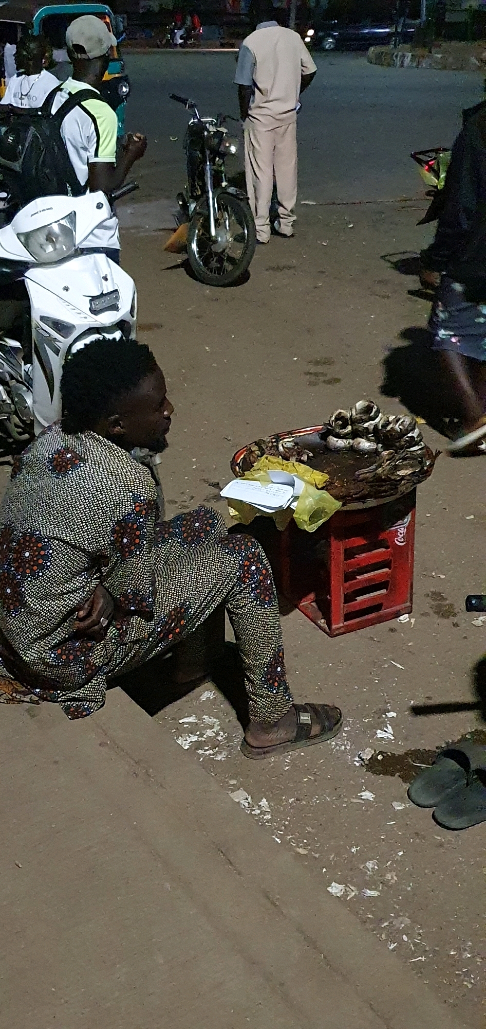 In Bukuru, Nigeria, a person sits on the ground next to a tray of food, likely fish, displayed on a red Coca-Cola crate. The individual is wearing a patterned fabric covering their body, with one hand resting on their lap, adorned with a ring. Their feet are clad in sandals. The background features several motorcycles, a white scooter, and other individuals in casual attire, suggesting a street scene or market area in the city. The lighting appears to be artificial, indicating it is either evening or night.