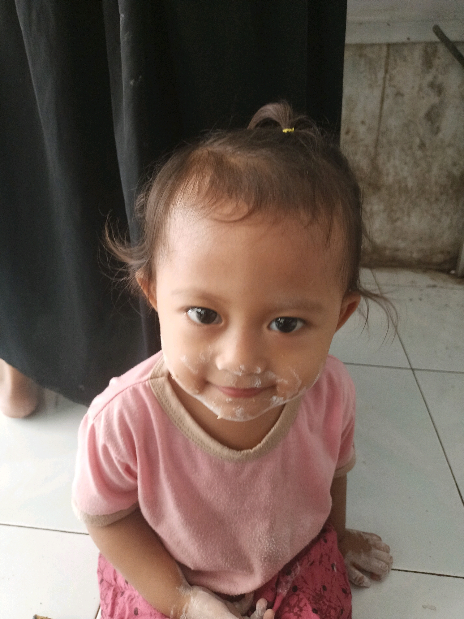 In Badas, Indonesia, a young child with a smear of white substance on their face sits on a tiled floor, looking directly at the camera. The child is wearing a pink t-shirt and red patterned pants. Their hair is pulled back with a yellow tie. In the background, a dark curtain hangs on the left, and a textured wall with some dirt marks is visible on the right. The floor tiles are light-colored and laid in a grid pattern. The child's hands are also covered in the white substance, suggesting they have been playing with it. The overall scene appears to be indoors, possibly in a home or domestic setting.