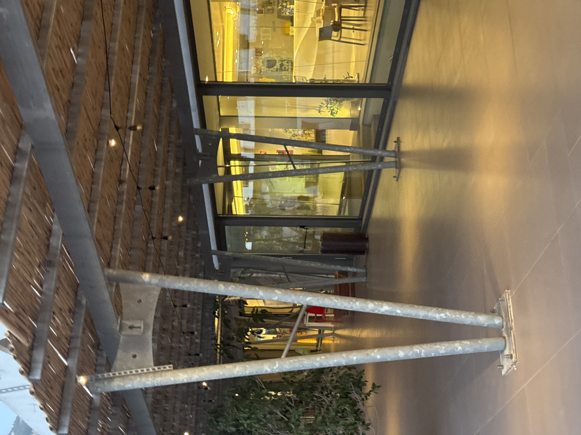 The image shows the exterior of a modern building with a large glass facade. The building appears to be a restaurant or cafe, with tables and chairs visible through the windows. Outside, there is a covered walkway with a slatted wooden roof supported by metal beams. String lights hang from the ceiling of the walkway, casting a warm glow. The time of day appears to be evening or night, as the interior lights are on and reflecting in the glass. The weather is likely mild, as people appear to be dining or socializing outdoors. The setting seems to be a pleasant, contemporary public space, possibly in a tourist area given the "Isola delle Femmine, Italy" context. There is no discernible text visible in the image.