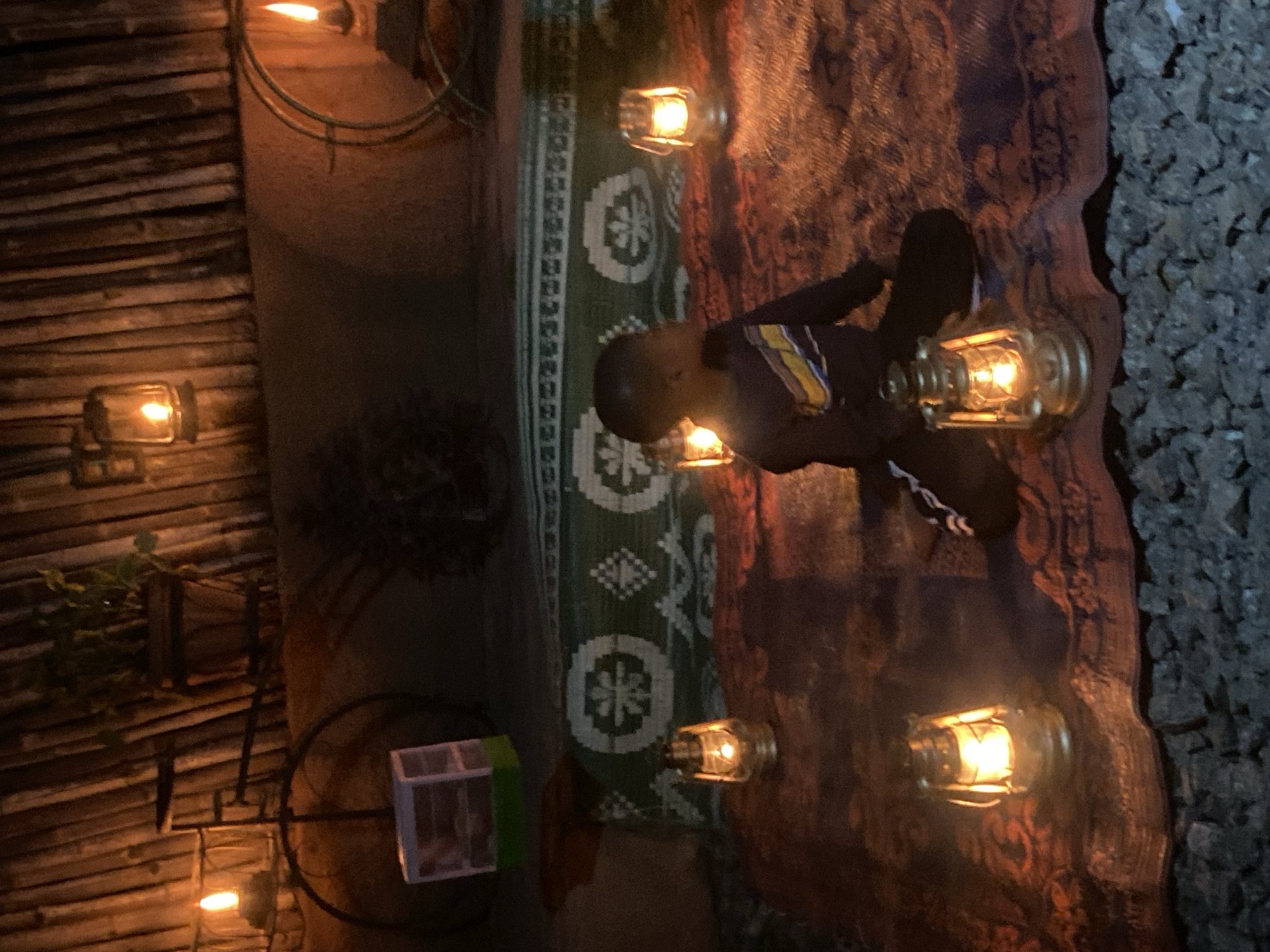 A nighttime view of an interior space in Barden Yana, Nigeria, is illuminated by multiple lanterns, casting a warm glow. The space is decorated with a patterned rug and a wooden wall. A person wearing a dark shirt with yellow and white stripes is present. The ground is covered with small rocks.