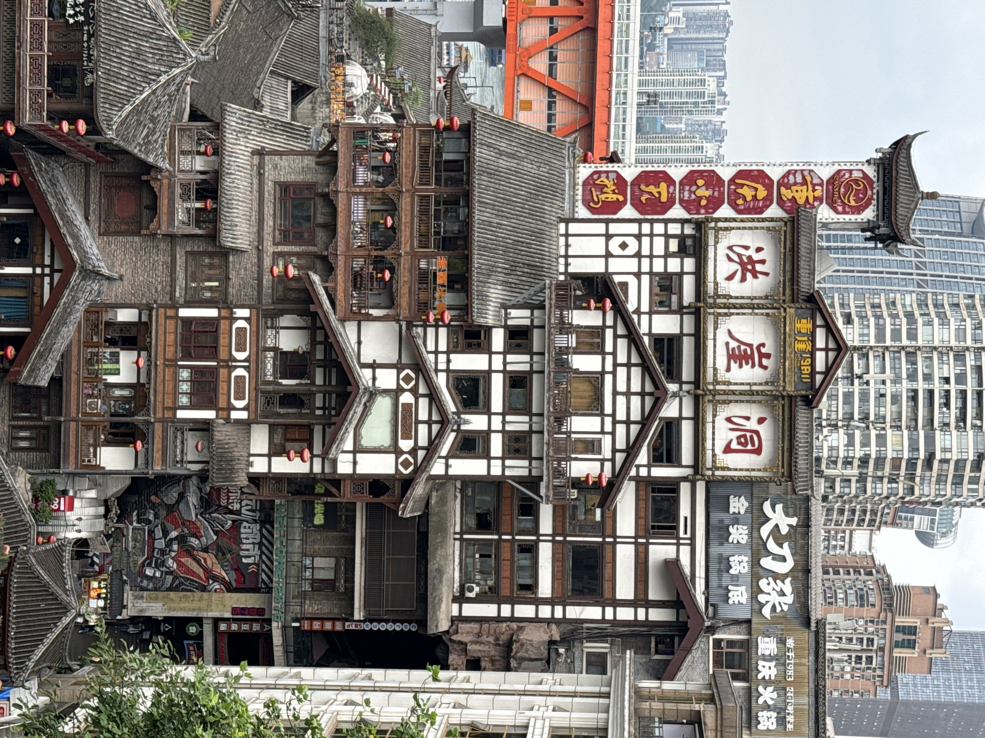 This is an eye-level, medium shot of the Hongyadong complex in Yuzhong District, Chongqing, China. The architecture is characterized by a mix of traditional Chinese and modern elements, featuring stacked buildings with intricate wood and white trim. Roofs are adorned with gray tiles, and numerous small red lanterns hang from the structures. Various signs with Chinese characters are displayed on the buildings. In the background, modern skyscrapers rise, contrasting with the traditional architecture of Hongyadong. An orange bridge structure can be seen connecting buildings in the mid-ground. Graffiti and other artwork are visible on the lower levels of the complex. Trees and greenery are present at the bottom of the frame. The sky is overcast and hazy.
