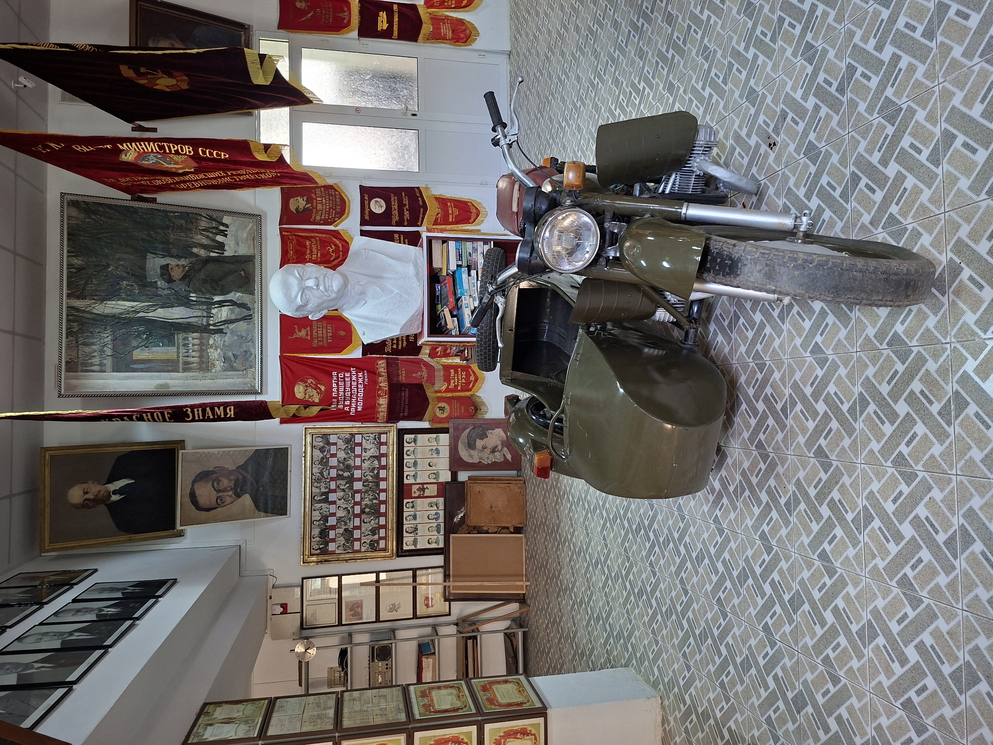 The photograph captures the interior of a museum or exhibition space in Ialoveni, Moldova. The room features walls adorned with framed portraits of historical figures, Soviet-era banners, and a large painting. In the foreground, a vintage olive-green motorcycle with a sidecar is parked. The floor is covered with a patterned tile mosaic. Among the exhibits visible are busts of Lenin, rows of framed photographs of individuals, and several shelves filled with books and what appear to be artifacts. The overall atmosphere suggests a historical display, likely related to Soviet or socialist history.