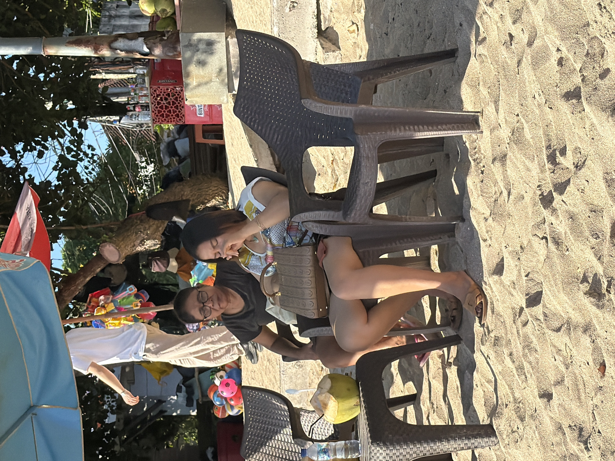 In Kuta, Indonesia, two women are seated on plastic chairs on a sandy beach, under a blue and red umbrella. The woman on the left, wearing glasses and a dark t-shirt, is looking down. The woman on the right, in a colorful top, is looking down and appears to be touching her face. A brown patterned handbag rests on her lap. To the left of the chairs, a green coconut with a straw and a bottle of water are on a small table. In the background, various items are displayed, including colorful balls, bottles on a crate, and some coconuts. A person in a white t-shirt and tan pants is standing behind the women. The scene depicts a relaxed atmosphere on the beach.