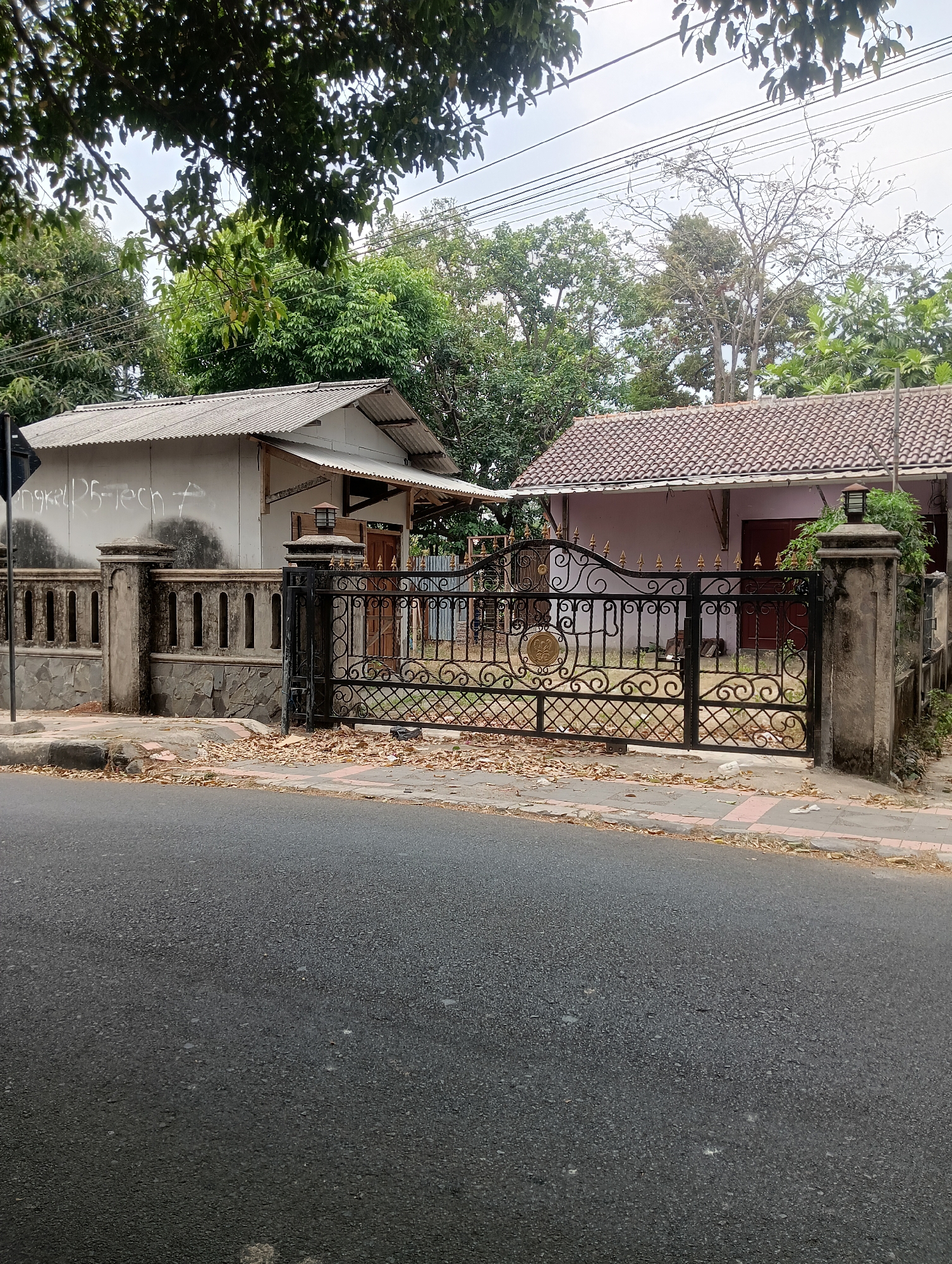 A wide shot captures a street scene in Ciporang, Indonesia, with two houses visible behind a decorative black metal gate and a stone and concrete fence. The house on the left has a corrugated metal roof and a white painted wall with graffiti. The house on the right has a terracotta tile roof and a light pink painted wall. A paved sidewalk runs in front of the houses, littered with fallen leaves. A paved road occupies the foreground of the image. The sky is overcast.