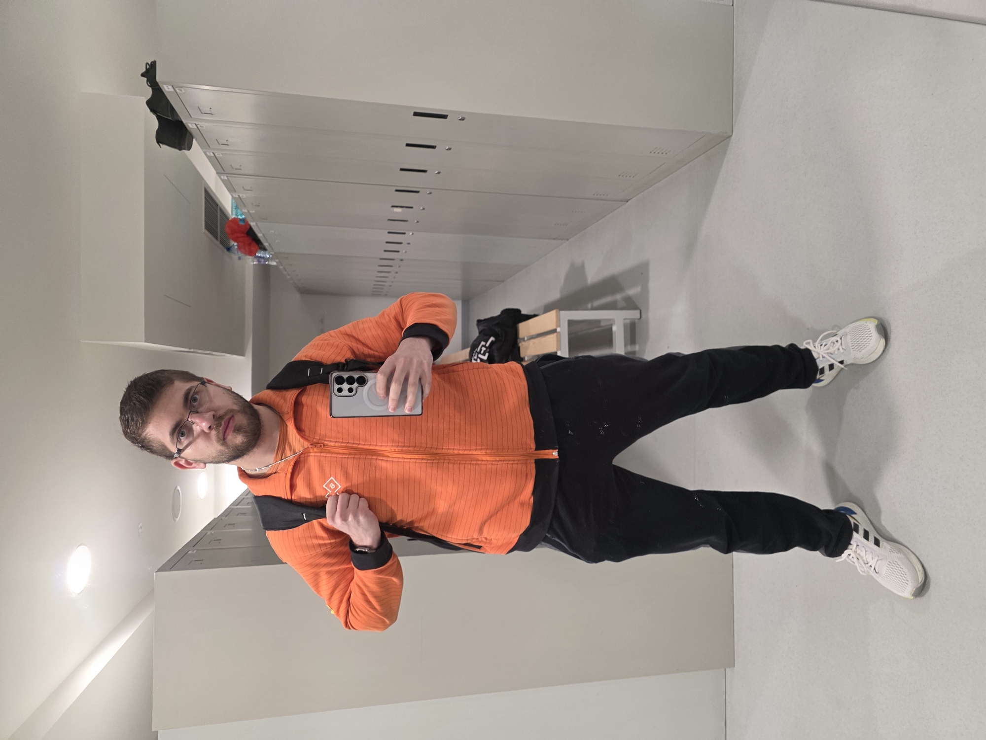 A man stands in a hallway with lockers and a bench, possibly a gym or a school facility. The man is wearing glasses, an orange jacket, black pants, and white athletic shoes. He is holding a smartphone with a metallic case in his hand, likely taking a selfie. The location is unlikely to be in Plovdiv, Bulgaria as there is no evidence to support this in the image. The hallway has gray lockers lining the wall and a gray floor. There is a bench in the background near the end of the hallway. The lighting is bright and appears to be artificial.