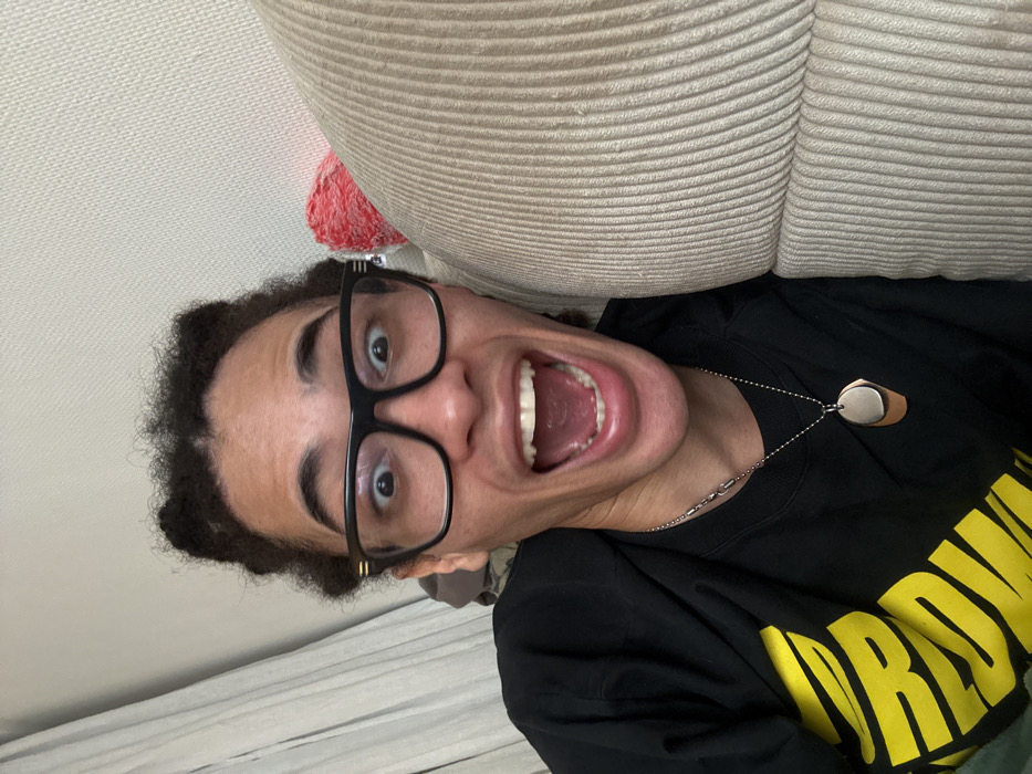 An individual is depicted in a close-up, indoor shot, presumed to be in Paris, France. The subject is a person with light-to-medium skin tone and dark, curly hair styled in two small knots on the top of the head. They wear black-framed eyeglasses with horizontal gold accents on the temples. Their eyes are wide open, and their mouth is open in a broad yell or laugh, conveying an exaggerated expression of excitement or surprise. The individual is wearing a black t-shirt with a partially visible yellow graphic, appearing to be stylized text. Two necklaces are visible: a silver-toned chain with elongated links and a ball chain with two overlapping, irregularly shaped pendants, one silver-toned and one copper-toned. The background on the left features a light-colored, textured wall with a light-colored curtain partially visible in the upper left. On the right, a significant portion of a light beige or tan ribbed couch is present, behind which a bright red object is partially obscured. The subject appears to be positioned directly adjacent to this couch.