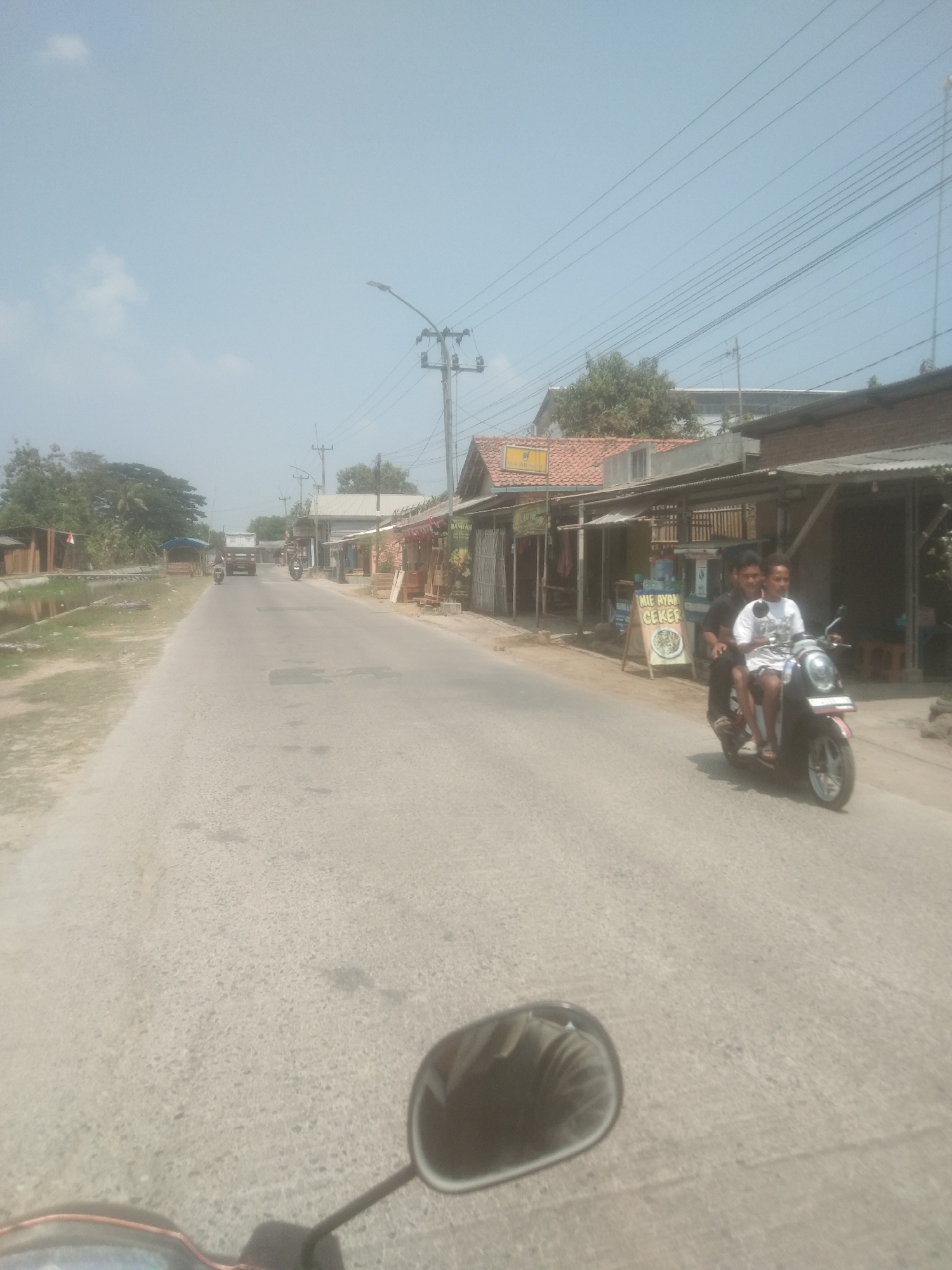 A clear, sunny day in Desa Sukra, Indonesia, shows a paved road with a slightly rough surface, bordered by grassy areas and small buildings on the right. Two young males are riding a dark-colored scooter on the right side of the road. The driver, wearing a black t-shirt, is looking forward, while the passenger, in a white t-shirt, is looking at the camera. A sign board with text "MIE AYAM CEKER" (Chicken Noodle Feet) is placed in front of a building on the right. In the background, a white truck is visible on the road, and a stream runs along the left side of the road, with some vegetation and simple structures bordering it. Power lines crisscross the sky. The image appears to be taken from another moving vehicle.