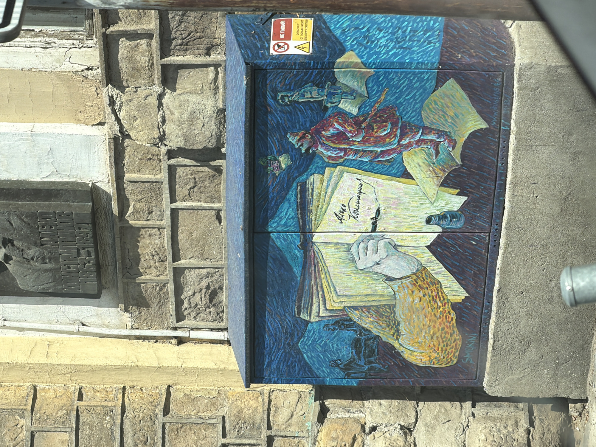 The media file displays an outdoor scene in Sofia, Bulgaria, featuring a vibrantly painted electrical box attached to an old stone building. The mural on the electrical box, rendered in a style reminiscent of Van Gogh with distinct, swirling brushstrokes and rich blue and yellow hues, depicts a literary theme. A hand, with a patterned sleeve, is shown holding a quill and writing in an open book. Several papers, some scattered, and an inkwell are present. In the upper section of the mural, a stylized, possibly reclining figure and what appears to be a rifle suggest a deeper, potentially historical or political context related to the writing. The cursive text "Алеко Константинов" (Aleko Konstantinov) is written on the book within the mural.
Adjacent to the electrical box, on the building's facade, is a dark memorial plaque bearing the engraved Cyrillic text "АЛЕКО КОНСТАНТИНОВ" and the years "1863-1897," identifying the subject as a historical figure, likely a writer or public figure. The building itself shows signs of age with rough stone blocks, worn plaster, and peeling paint. A yellow warning sign is affixed to the top of the electrical box, with Cyrillic text that reads: "НЕ ПИПАЙ!" (Do not touch!), and "ОПАСНОСТ ОТ ПОРАЖЕНИЕ ОТ ЕЛЕКТРИЧЕСКИ ТОК" (Danger from damage from electric current). A signature "SMOKINI" is visible on the bottom right of the mural, and another partial signature above it reads "Mimi Karadzhova 8.6." and "1983/93". The scene is well-lit, indicating daytime.