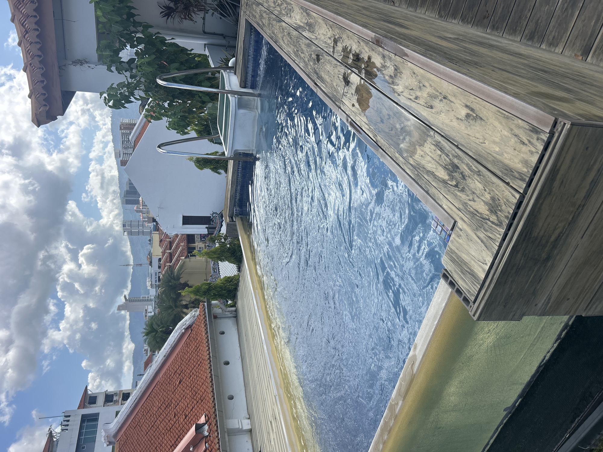 A rooftop swimming pool is pictured in George Town, Malaysia, with the cityscape and mountains visible in the background under a partly cloudy sky. The pool's water is a clear blue, with ripples indicating movement. The pool's edge is lined with wooden decking and a white structure with metal railings, presumably a Jacuzzi or spa feature. In the foreground, the textured wooden deck is visible, with its planks showing a natural wood grain pattern. Beyond the pool, traditional terracotta-tiled roofs of older buildings in George Town are prominent, contrasted with modern high-rise buildings in the distance. Green cypress-like trees and lush foliage add a touch of nature to the urban landscape. The overall impression is a serene and luxurious amenity set against a vibrant and historic city backdrop.