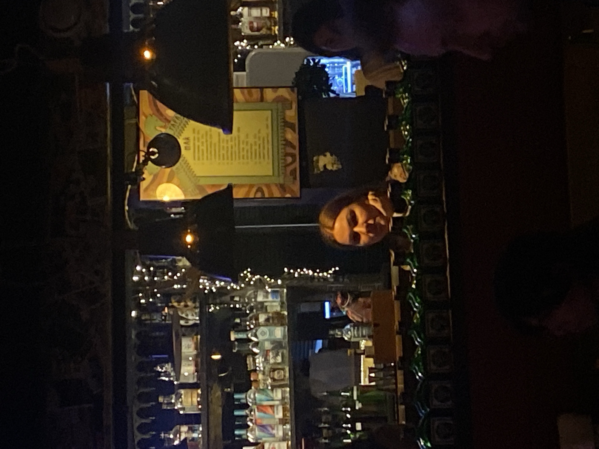 An indoor scene, likely a bar or dimly lit restaurant, is captured at night. The image is oriented vertically with the top shifted to the right. A prominent individual, appearing to be a woman, looks directly at the camera from the mid-right. Her face is well-lit, while other patrons are visible in the background, largely obscured or out of focus, including a head in the upper right and another in the lower right.
The background features a bustling bar area on the left, adorned with numerous bottles of alcohol and decorative string lights that cast a warm glow. Behind the prominent person, a framed sign or menu hangs on the wall. The frame has a light green border and warm-toned, abstract patterns. Inside, a light-colored rectangle displays text, which appears to be a list. Visible larger text reads "ТАРЕЛКА" (TARELKA) and "MAP" (MAP) at the top. A black, circular lamp or microphone with a glowing center is positioned to the left of this sign. Further in the background, a blue-lit object resembling an aquarium or display case can be seen. Along the lower edge of what might be a bar counter or partition, there's a decorative border of dark squares interspersed with green, vine-like garlands. The overall atmosphere is social and intimate, characterized by the low light and decorative elements.