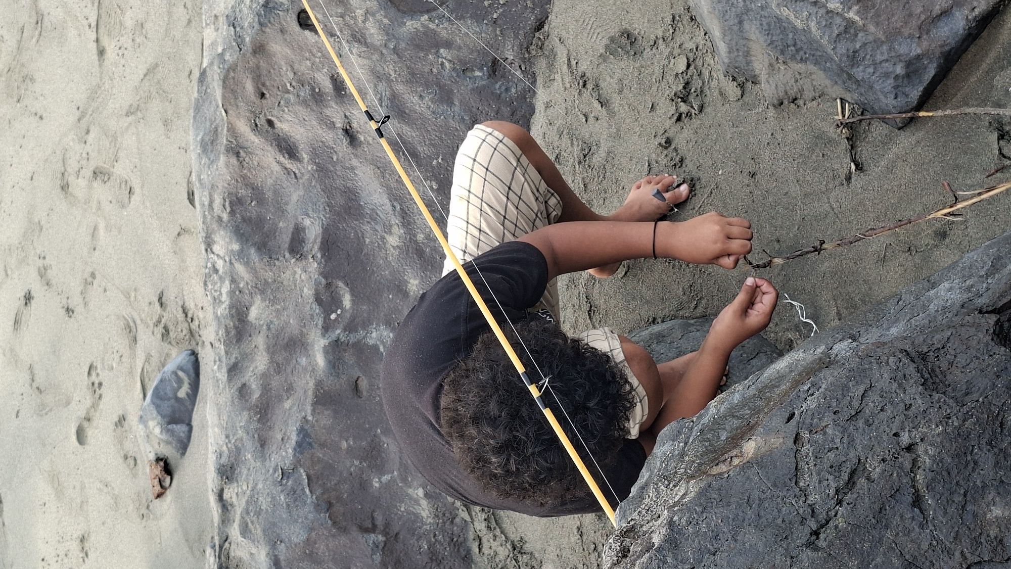 A person is crouched on a sandy, rocky shoreline, attempting to tie something to a thin branch. The person, wearing a black t-shirt and checkered shorts, has dark curly hair and is wearing a black bracelet on their left wrist. Their bare feet are visible in the sand, and they are wearing flip-flops. A yellow fishing rod is propped up behind them, with its line extending towards the person's hands. The environment appears to be a beach with large, dark rocks and fine, light-colored sand. There are footprints visible in the sand. The scene is likely Lhokseumawe, Indonesia, given the typical coastal landscape.