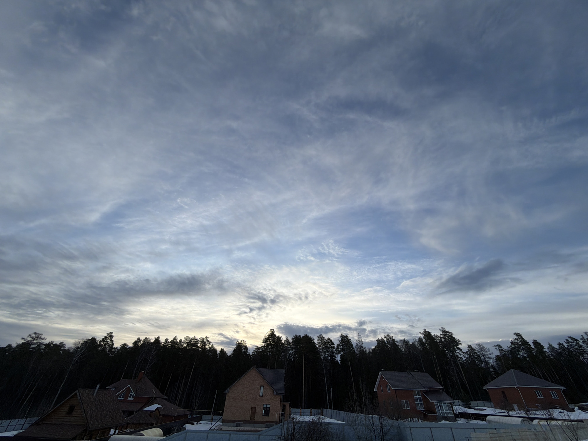 The image displays a suburban or rural landscape in Chaikovsky, Russia, under a partly cloudy sky. The upper two-thirds of the frame show a expansive sky with a gradient from light blue to a darker grey-blue, filled with white and grey wispy cirrocumulus and altocumulus clouds, with a brighter, almost golden-white area visible near the horizon. Below this, a dense, dark line of coniferous trees, primarily pines, forms the middle ground, silhouetted against the sky. Several detached residential structures are visible below the tree line. On the far left, a log cabin-style house with a dark roof is partially obscured. Towards the center-left, a two-story light brown brick house with a dark grey gabled roof and a single vertical window on the second floor stands prominent. Further to the right, two additional red brick houses with dark grey gabled roofs and multiple windows are present, with one appearing to be under construction or recently completed. The ground in the foreground and immediately surrounding the houses is partially covered in snow, with areas of exposed earth and various temporary fencing or barriers. No individuals or active subjects are present within the scene.