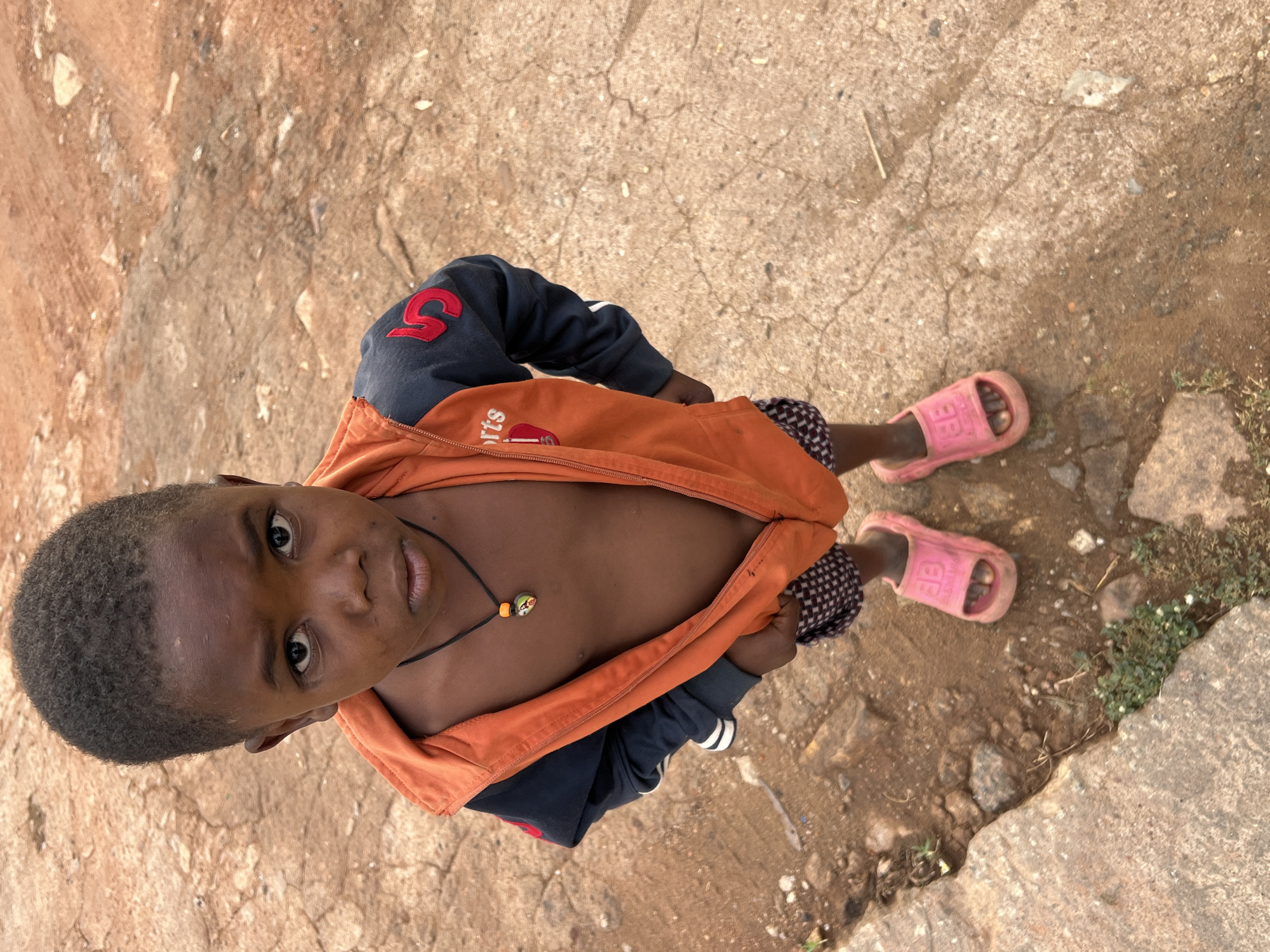 An overhead, high-angle shot captures a young child standing outdoors on a rough, earthy ground. The child, with dark skin and short, dark hair, looks directly up at the camera with a neutral and attentive expression.
The child is wearing a dark blue long-sleeved top with a prominent red number '5' on the left sleeve. Over this, an orange zipped jacket is worn partially open, revealing the child's bare chest. Faint text, "rts," and a small red car logo are visible on the left side of the orange jacket. A black string necklace with a small, colorful, spherical pendant hangs around the child's neck. Patterned shorts or trousers are visible beneath the jacket, and the child wears pink plastic slide sandals, with the letters "BB" embossed on the strap of the left sandal.
The background consists of dry, cracked dirt and small stones, along with scattered patches of sparse green vegetation, indicating an unpaved, outdoor setting. The bright, even lighting suggests it is daytime. The scene conveys a candid portrait of the child in what appears to be a warm, possibly rural or semi-urban environment in Gura Top, Nigeria.