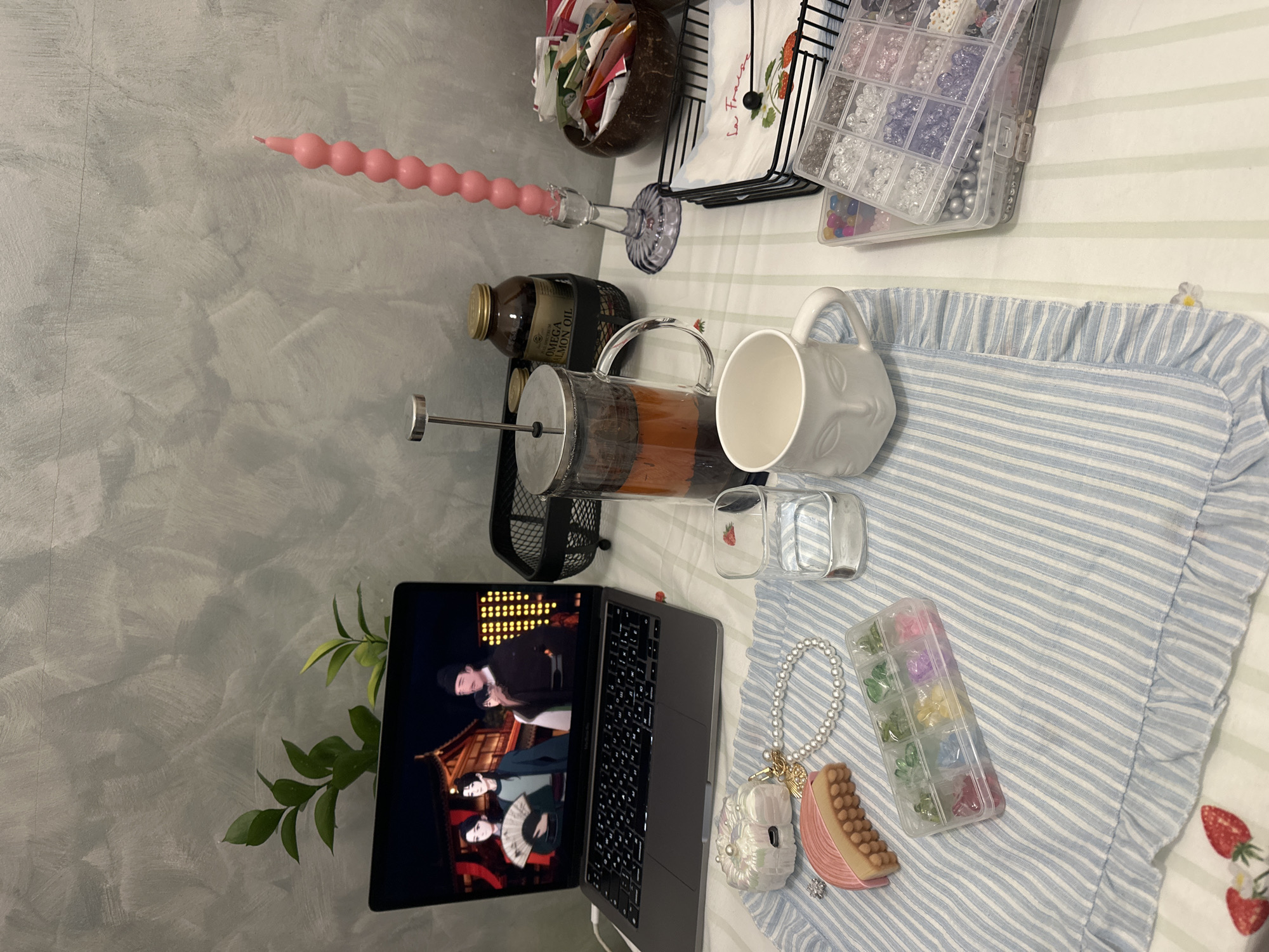 The scene is an indoor setting in Šušanj, Montenegro, featuring a detailed arrangement of objects on a table, likely a desk. The background consists of a light grey wall with a textured, stucco-like finish. The table is covered by a light-colored cloth with faint vertical green stripes, partially overlaid by a blue and white striped fabric with a ruffled edge.
On the table, a silver-colored laptop is open and powered on, displaying an animated scene with four characters—three adults and one child—dressed in traditional East Asian attire, set against a backdrop of traditional East Asian architecture at night. Behind the laptop, a small green plant with two distinct stems is visible.
To the right of the laptop, a clear glass French press contains a dark reddish-brown liquid. Further right, a clear square glass holds water, and beside it is an empty white ceramic mug featuring a stylized human face in relief. A tall, slender pink candle, composed of multiple spherical segments, stands upright in a clear, fluted glass candlestick holder. Behind the candle, a brown glass jar labeled "OMEGA SALMON OIL" is placed within a small black wire basket. A coconut shell bowl filled with various colorful sachets or packets is situated to the right of the candle. Another black wire basket holds folded white napkins adorned with strawberry motifs and the text "La Fraise."
In the foreground, on the blue and white striped fabric, a white, intricately textured case (possibly for AirPods) decorated with a pearl and a gold-colored charm is present. Next to it, a string of white pearl beads, resembling a bracelet, is laid out. A pink, ribbed, rectangular object, possibly a decorative item or soap, lies near the pearls. A small, translucent plastic organizer box with nine compartments holds colorful gummy bear-shaped beads (green, yellow, purple, pink). Further to the right, a stacked group of larger, clear plastic organizer boxes contains an assortment of beads of various colors, shapes, and sizes, including clear, white, grey, and pastel spherical beads, some appearing pearlescent. A small artificial strawberry decoration is positioned on the bottom left corner of the blue and white striped fabric. The overall arrangement suggests a personal leisure or crafting activity.