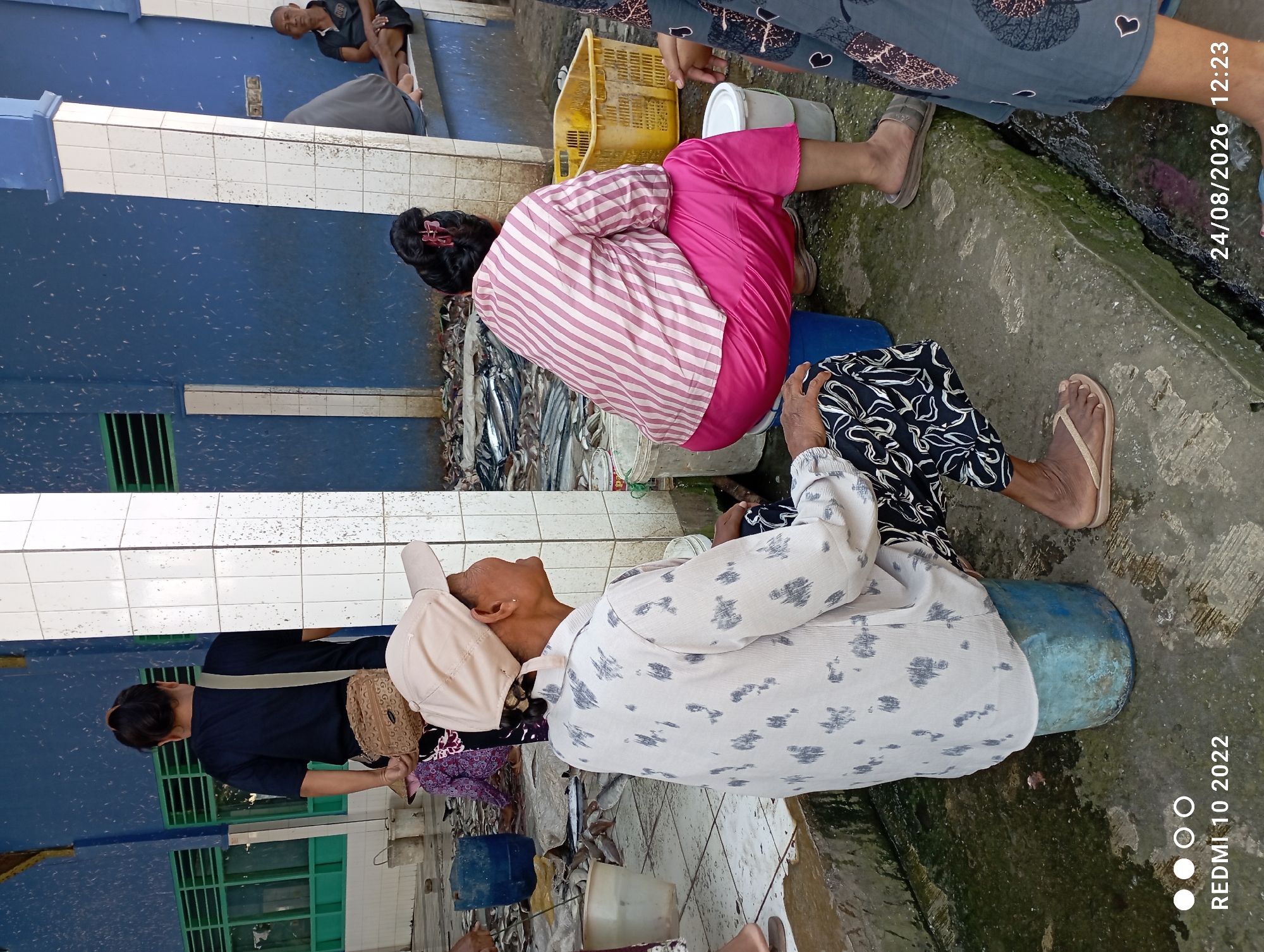 The image depicts an outdoor market scene, likely a fish market, in a location with blue walls and white tiled pillars. In the foreground, two women are seated on buckets. The woman on the left wears a light-colored, patterned shirt and black and white patterned pants, with a light-colored cap. The woman on the right is dressed in a pink and white striped shirt and bright pink pants. A yellow plastic basket is visible behind her. In the background, a man in a dark shirt is visible, and other people are present. The ground is wet and appears to be concrete, with many dead fish scattered around, indicating a recent sale or processing of fish. There are also several buckets and containers scattered throughout the scene. The overall environment appears somewhat cluttered and utilitarian, typical of a working market.