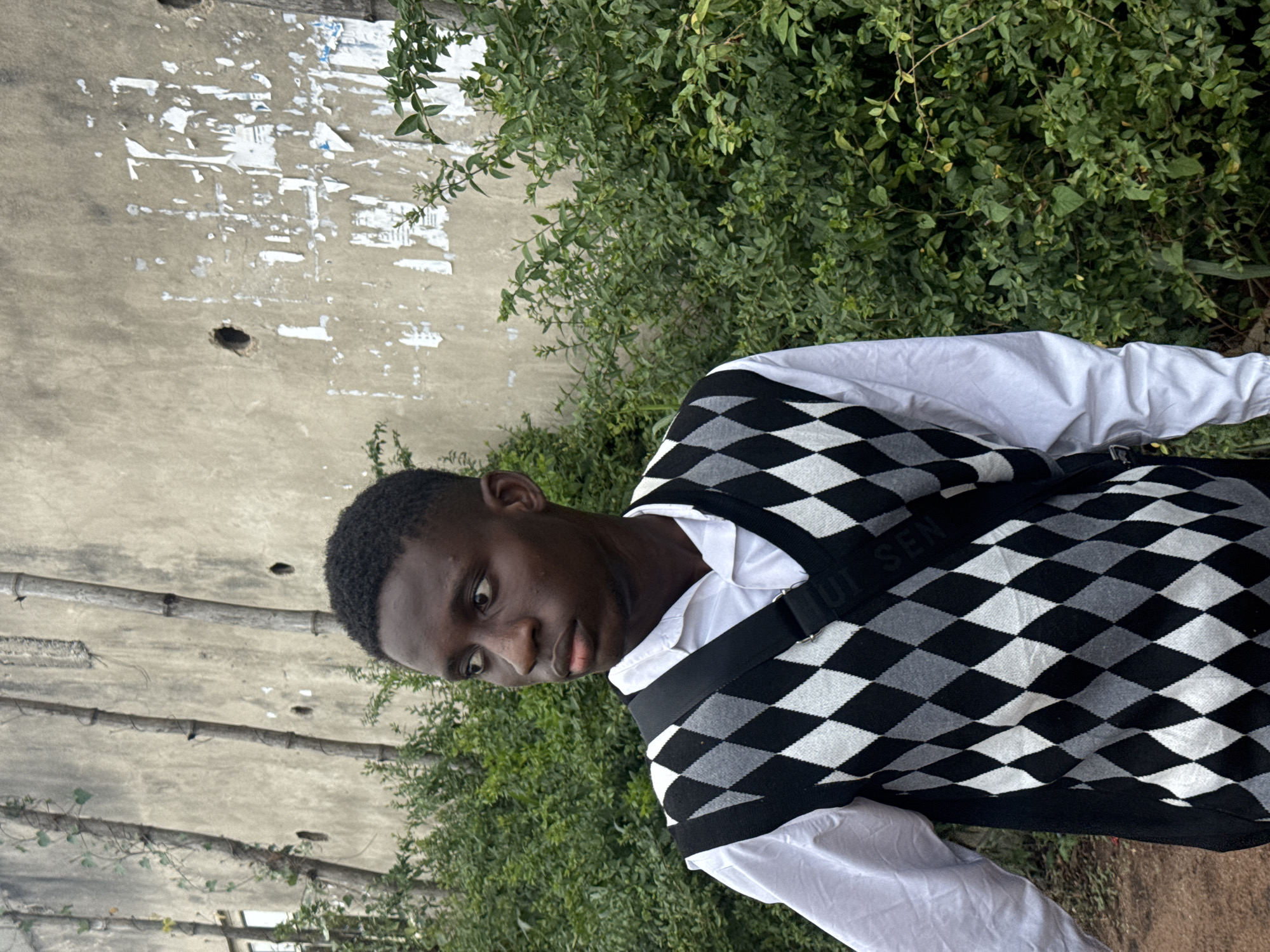 An outdoor scene in Ihiagwa, Nigeria, features a young male subject positioned slightly off-center to the right, facing and gazing towards the left. The individual has dark skin and short, dark hair. He is wearing a white long-sleeved collared shirt beneath a sleeveless V-neck argyle sweater vest patterned in black, white, and gray diamonds. A black strap from a bag, bearing white text partially visible as "UI SEN," crosses diagonally over his chest. The background consists of a weathered, light-colored concrete wall displaying several dark, circular holes and multiple remnants of torn white paper or posters. Three slender wooden poles, possibly bamboo, are vertically arranged against the wall, with one pole showing minimal vine growth. Dense green bushes and foliage surround the subject, obscuring the lower portion of the wall. A white-framed window is partially visible on the far left, behind the wall. The ground in the lower-left foreground is reddish-brown earth.