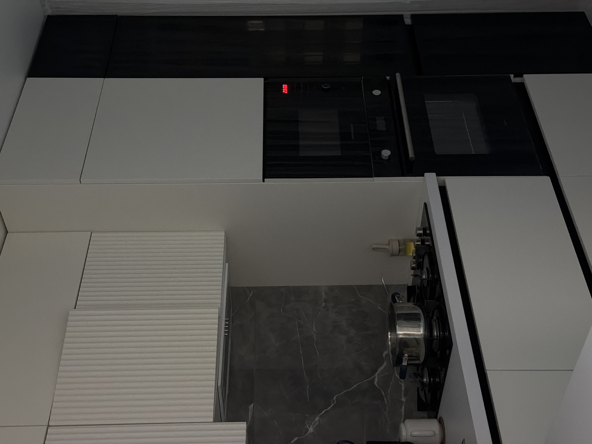 This is a wide, eye-level, interior shot of a modern kitchen, likely in Naberezhnye Chelny, Russia. The kitchen features a dual-tone design with white upper cabinets and a black appliance wall. The white cabinets have a ribbed texture and are mounted above a dark marble backsplash and a gas stovetop. A stainless steel pot sits on the stovetop, suggesting recent or ongoing cooking. Below the stovetop, white cabinets with matte finishes line the counter. To the right, a tall black unit houses a microwave and an oven, with a red digital display showing "23:00". The overall impression is one of clean lines and contemporary design, with a focus on functionality.