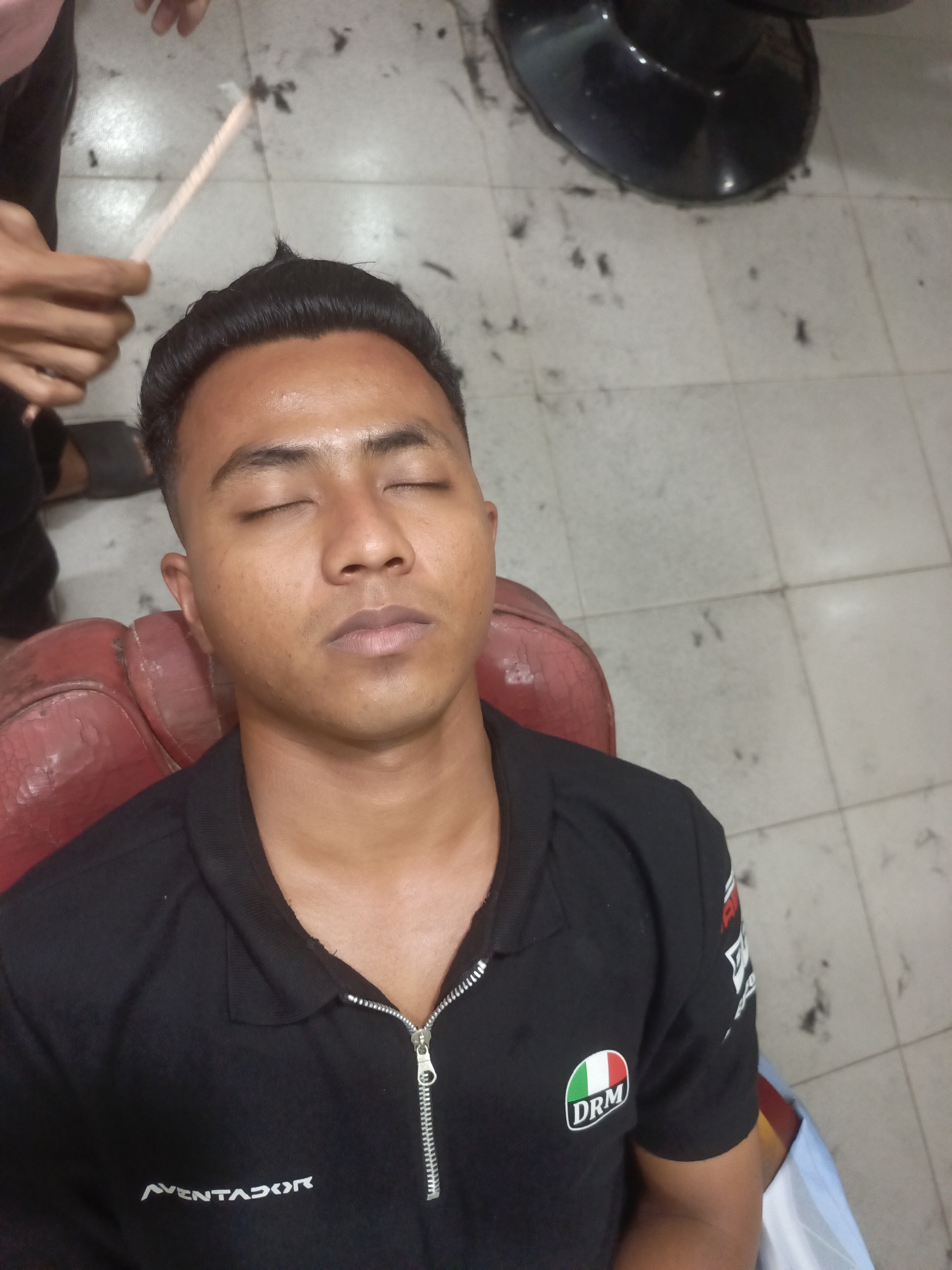 A young man with closed eyes, likely in his early twenties, is seated in a barber chair, undergoing a haircut. His head is tilted slightly upwards. He is wearing a black polo shirt with a zipper and an emblem on the left chest featuring the Italian flag colors and "DRM" written in white. The floor, made of light-colored square tiles, is littered with small clippings of hair, indicating the ongoing haircut. A dark, reflective object, possibly a salon chair base, is visible in the upper right corner. In the upper left, a person's hands are visible, holding a thin, light-colored stick-like object, presumably a comb, near the man's head. The overall scene suggests a typical barbershop environment. This image was taken in Lhokseumawe, Indonesia.