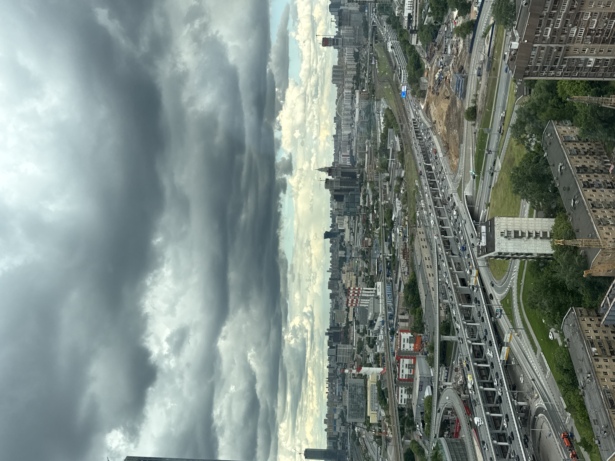 The image presents an elevated view of Khimki, Russia, under a sky dominated by heavy, gray storm clouds on the left and patches of bright clouds on the right. The cityscape includes a dense network of railways, highways with active traffic, and various buildings ranging from mid-rise apartment complexes to taller structures resembling industrial or commercial sites. Visible construction work is present alongside the railways. Buildings include both residential and industrial types. The overall environment suggests a typical urban setting with elements of development, transportation, and potential weather disturbances. No people are directly visible.