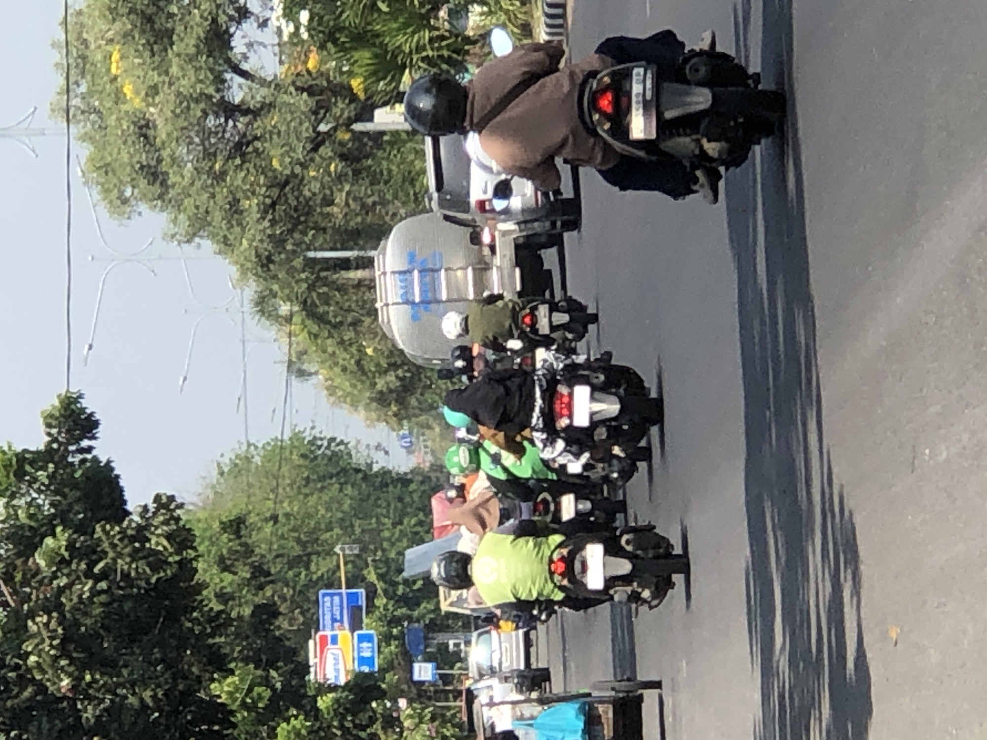 The image depicts a street scene in Larangan, Indonesia, on a sunny day. Several people are riding motorcycles on a paved road lined with trees. In the foreground, a person wearing a brown shirt and a black helmet is riding a motorcycle. Behind them, another person in a bright green vest and a black helmet is also on a motorcycle. Further back, a group of motorcyclists are visible, some wearing helmets and others not. A large white tanker truck is in the middle ground, along with a silver SUV. Traffic lights and street signs are visible on the left side of the road, including a blue sign with white text. The environment suggests a typical urban or suburban street with moderate traffic.