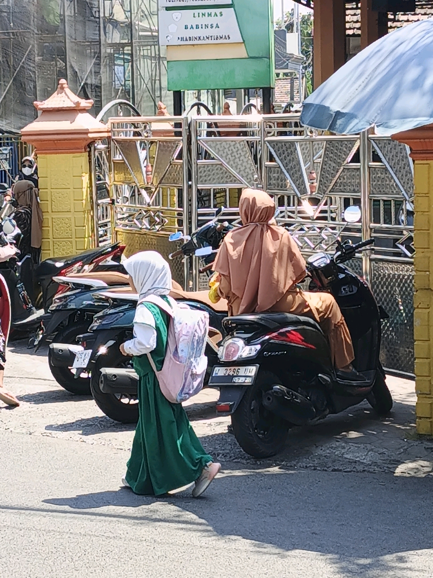 In Ketegan, Indonesia, a young girl wearing a white hijab, green dress, and pink backpack walks away from the camera, past several parked motorcycles. She is walking on a paved surface in front of a decorative yellow wall and a metal gate. Behind the motorcycles, a person in a tan hijab and pants sits on a black scooter. A blue umbrella is visible to the right of the frame. A sign with green and white panels is mounted on a pole in the background.
