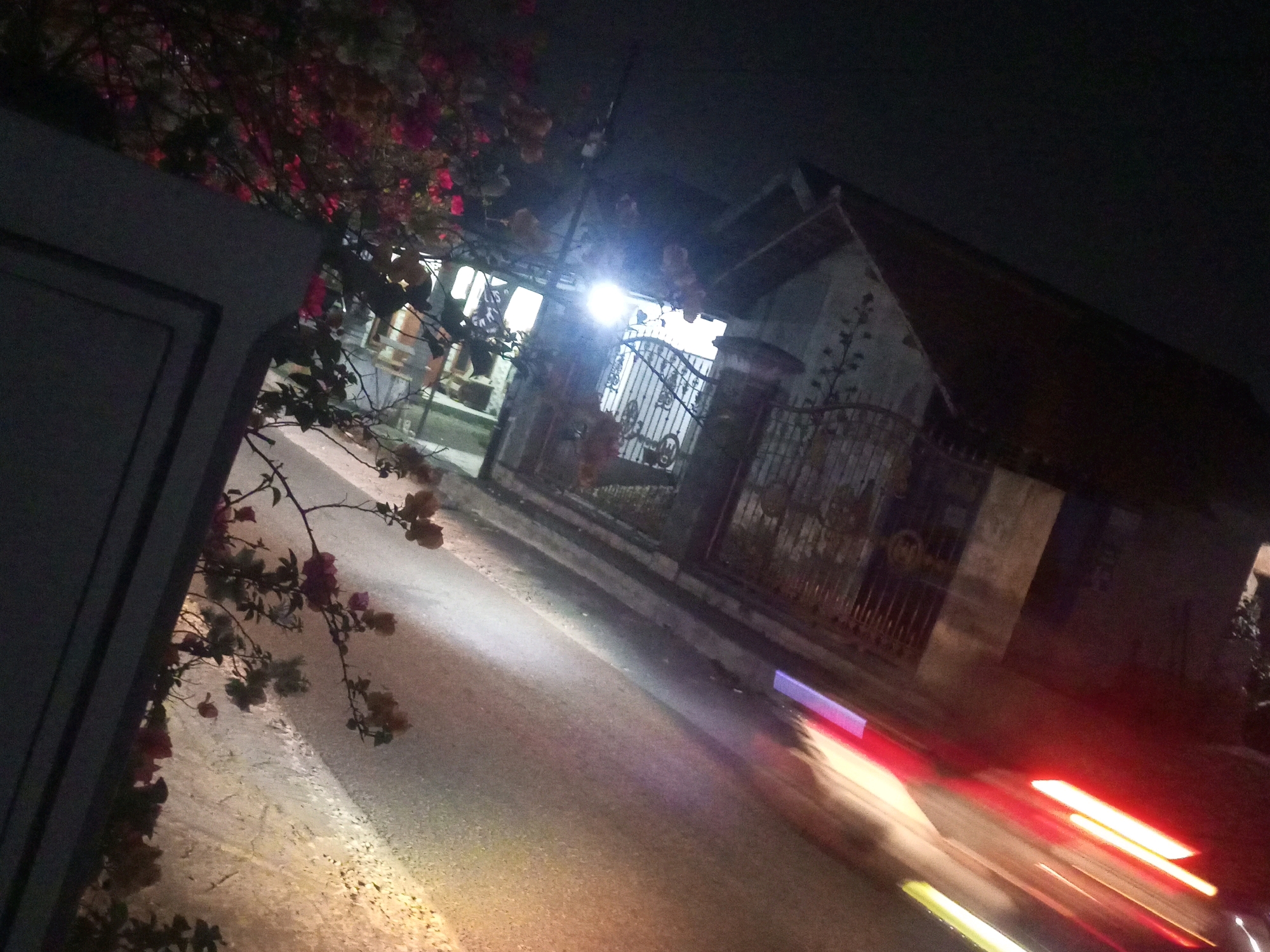 In the city of Ngasem, Indonesia, a street scene unfolds at night. A dimly lit house with a pitched roof stands behind a metal gate, illuminated by a bright security light. The house appears to have a light-colored wall and a dark roof. The gate is intricate with vertical bars and decorative elements. In the foreground, branches of a tree with small pink and orange flowers hang over a dark, textured wall. The street in front of the house is paved and wet, reflecting the lights from passing vehicles. Streaks of red and white light indicate the movement of cars, suggesting a dynamic nighttime environment. The overall impression is one of a residential street captured during the night, with a focus on the interplay of light and shadow.