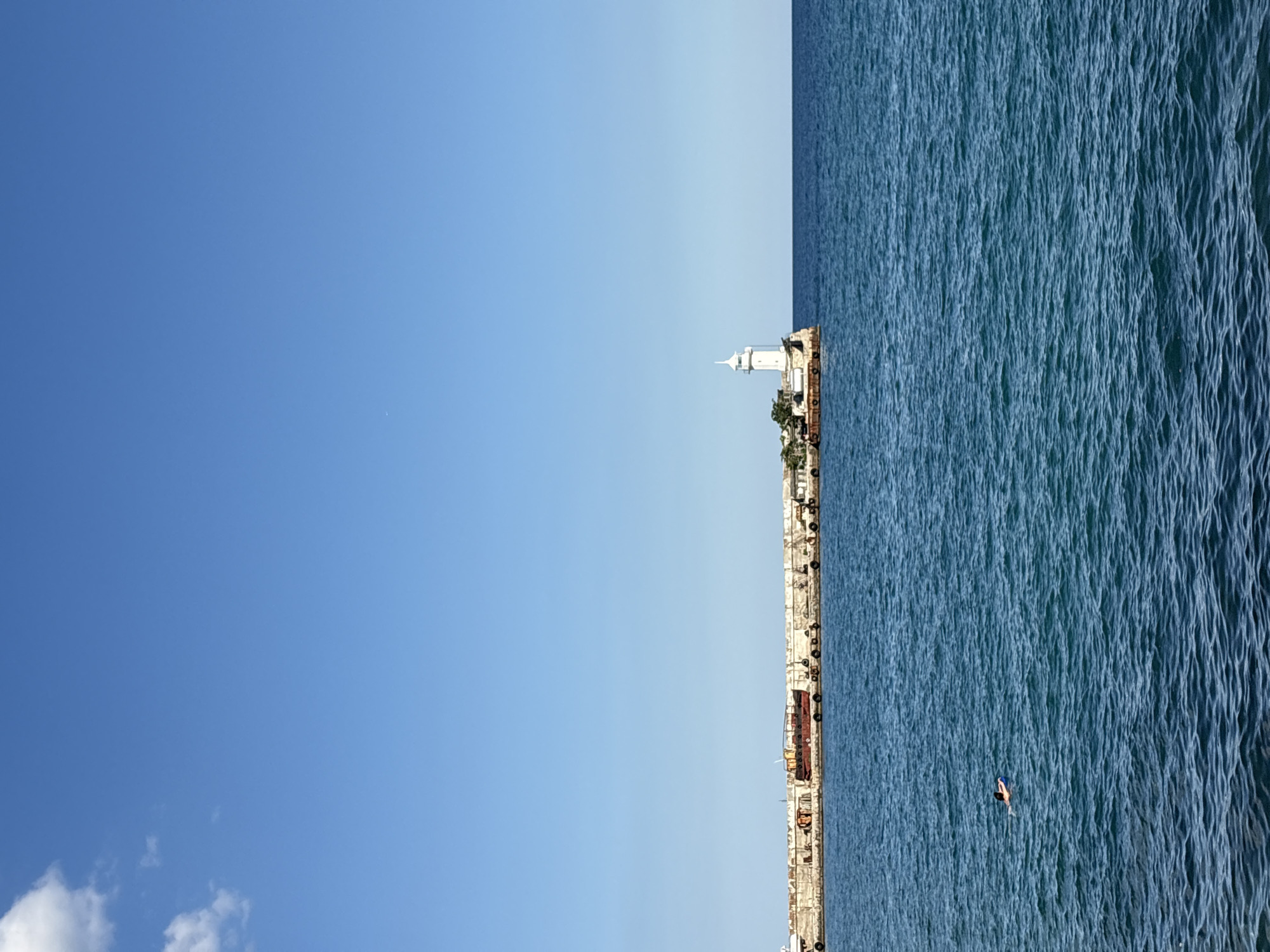 A vertical, eye-level shot shows a bright, cloudless blue sky dominating the top two-thirds of the frame. Below, the deep blue sea stretches to the horizon, meeting a breakwater that juts out from the left side of the image. The breakwater is constructed of rough concrete blocks and is lined with industrial elements, including rusted metal structures, a red barge, and numerous black tires used as fenders. On the far right of the breakwater stands a white lighthouse with a conical roof, its base appearing to be a concrete platform. In the middle distance of the water, a person is swimming with a blue float. The scene is set in the city of Yalta, Ukraine.
