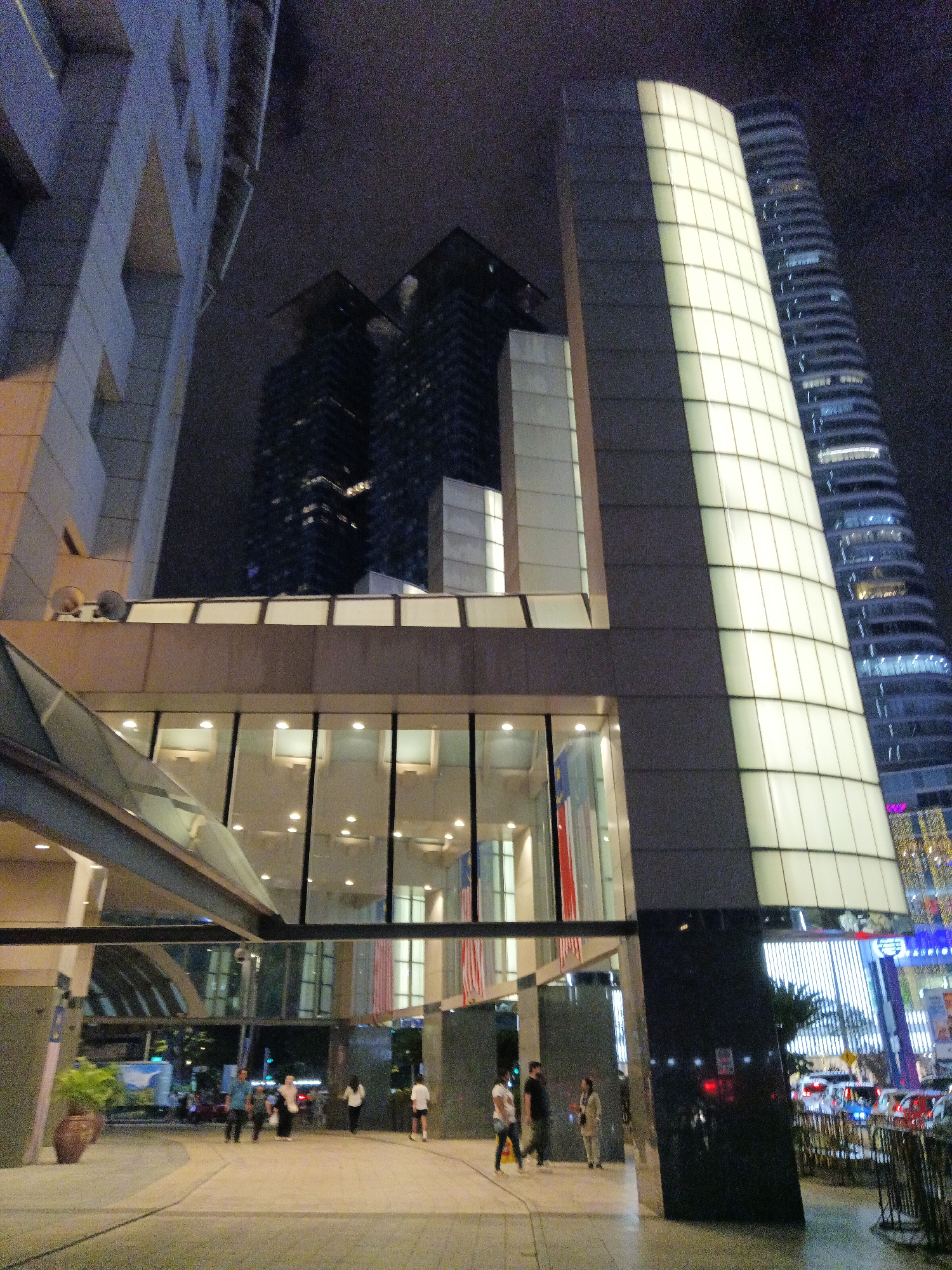 The image depicts a nighttime view of a modern urban environment in Kuala Lumpur, Malaysia. The foreground features a pedestrian walkway with several people walking and standing. To the left, a large potted plant is visible. The middle ground is dominated by the facade of a building with large glass windows, reflecting the lights and a covered walkway. Malaysian flags are displayed vertically near the entrance.
In the background, several contemporary skyscrapers rise into the night sky. One prominent building features a curved facade with a grid of illuminated panels, appearing to be a large screen or a lit architectural element. Another skyscraper to its right has a distinctive wavy or ribbed design, also illuminated with blue lights. The overall scene is characterized by the architecture of modern buildings, artificial lighting, and the presence of people in an urban setting at night. The weather appears to be dark and possibly overcast, contributing to the nighttime ambiance.