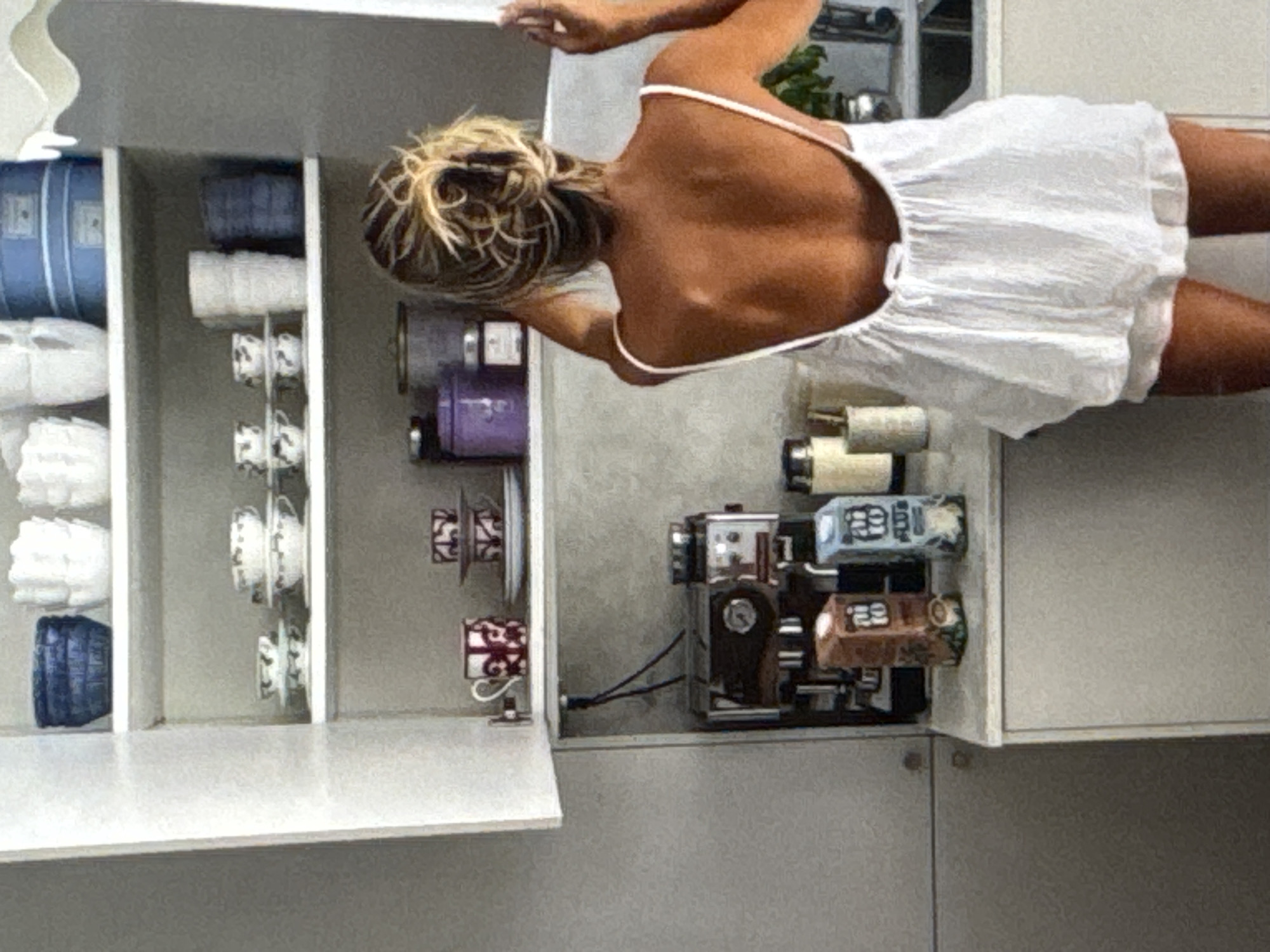 In Kuta Selatan, Indonesia, a woman with sun-kissed skin and her hair pulled back in a messy bun is facing away from the camera, standing in front of an open white cabinet filled with kitchenware. She is wearing a white, loose-fitting dress with spaghetti straps and a low back. The cabinet shelves display various items, including blue and white ceramic bowls, stacks of white mugs, and two blue containers on the top shelf. The middle shelf holds a collection of small cups and saucers, white ceramic cups, and a dark blue container. On the bottom shelf, a red and white patterned mug sits next to a stack of plates. Below the cabinet, a coffee machine is visible, along with two small cardboard cartons, one brown and one blue. The woman's hands are raised, adjusting her hair. The background suggests a kitchen setting with a sink and a countertop. The lighting is soft and natural.