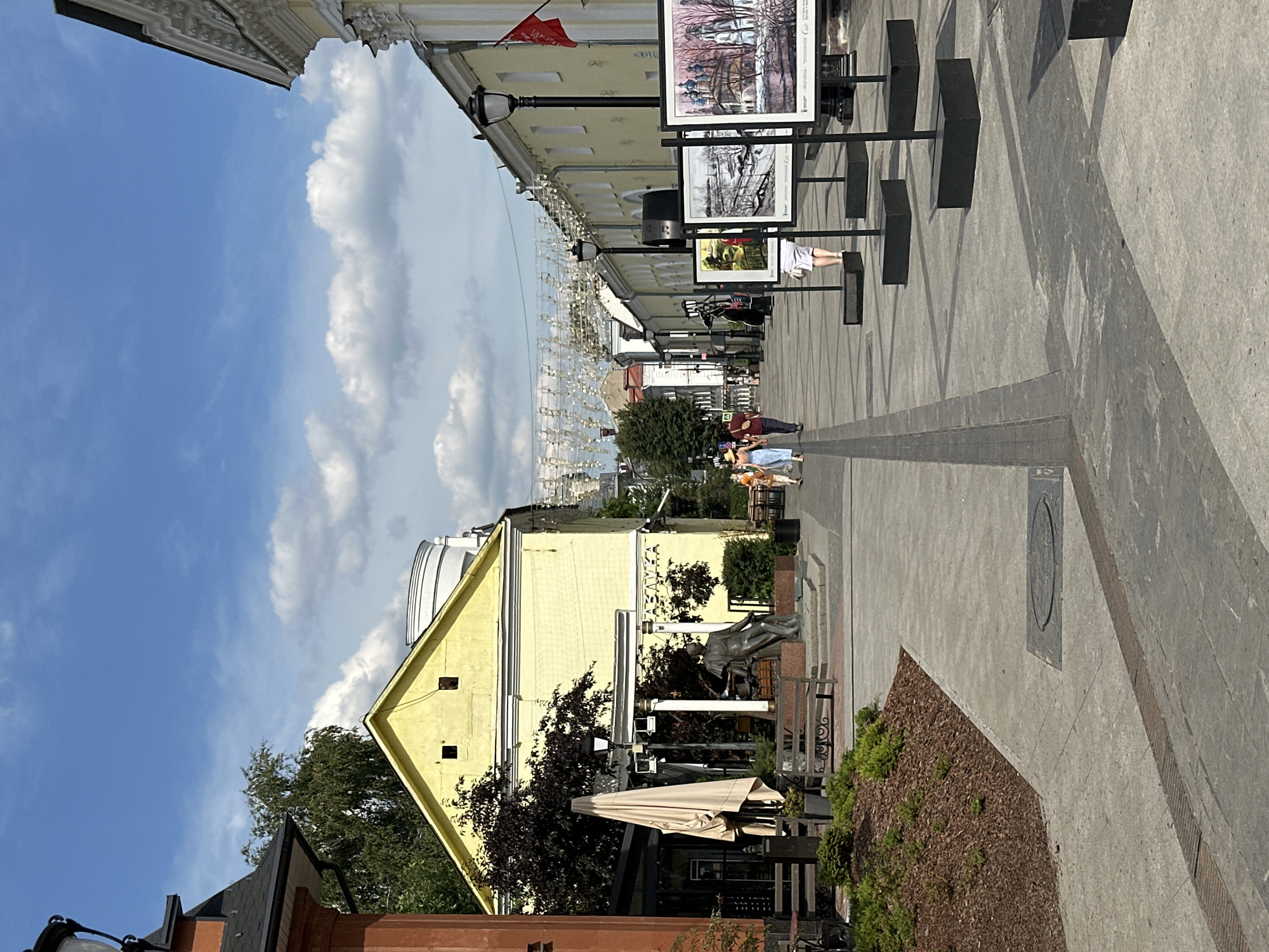 The image depicts a sunny day on a pedestrian street in Putyatino (Semivragovskaya S/A), Russia. The street is lined with buildings, one a bright yellow structure on the left and a longer, pale yellow building with white trim on the right. Decorative garlands of what appear to be musical notes are strung across the street overhead.
In the foreground, on the left side, is a statue of a person seated on a bench, surrounded by landscaping. Adjacent to this is a covered outdoor seating area with tables and chairs, and a large, light-colored umbrella.
The pavement is made of large, gray rectangular stones. A black metal grate runs along the edge of the sidewalk on the right side.
Several large framed artworks are displayed on black stands along the right side of the street. These artworks appear to be paintings of landscapes and architecture.
A few people are walking along the street. On the right side, a person wearing a white skirt and top is looking at the artwork. Further down the street, a group of three people, two adults and a child, are walking away from the camera. On the far left, two figures are visible in the background.
The sky is blue with scattered white clouds. Trees with dark green foliage are visible in the background, particularly behind the yellow building.