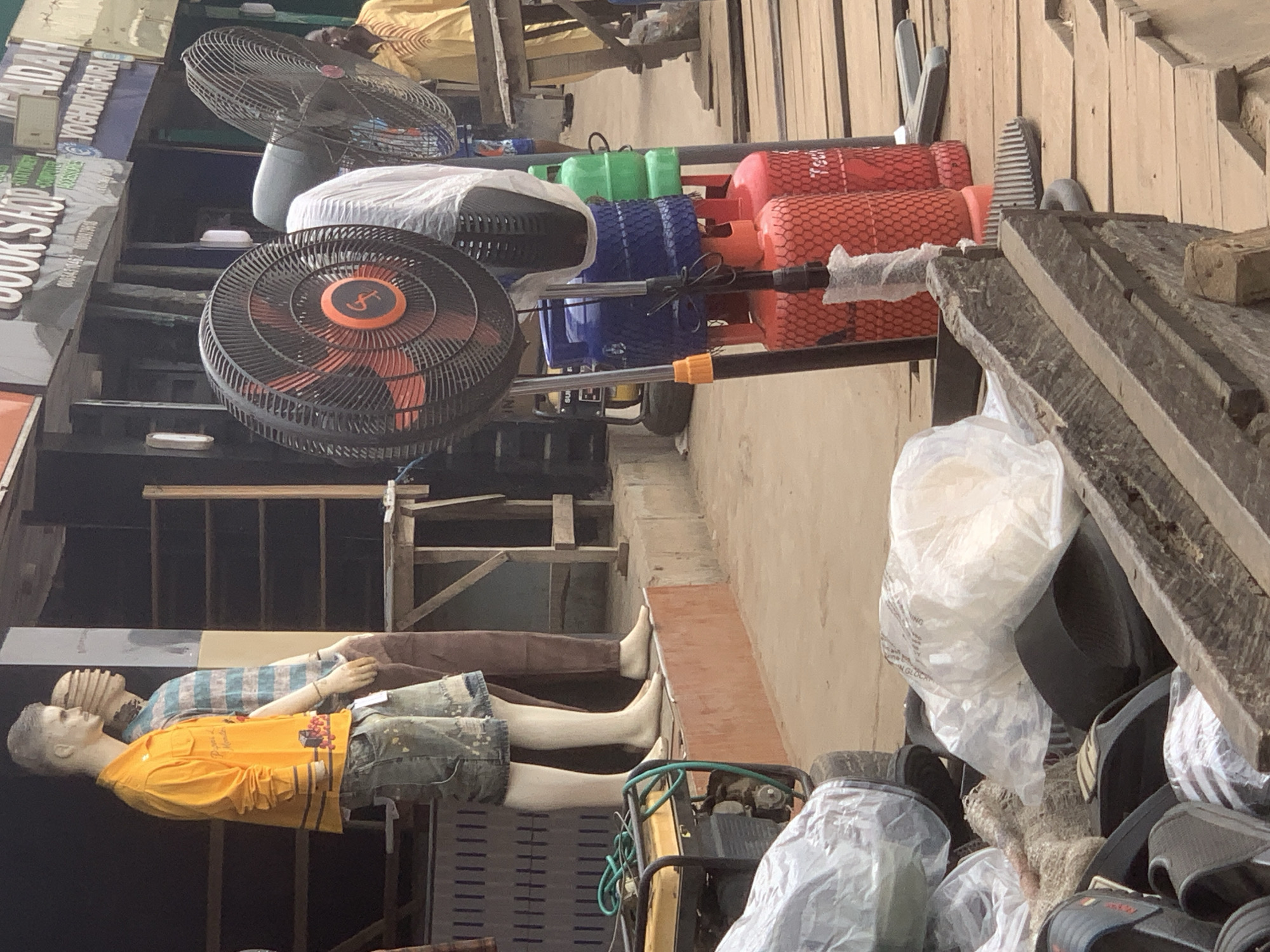The image depicts an outdoor market scene in Shagamu, Nigeria, characterized by a collection of merchandise and commercial activity. In the foreground, a pile of black sandals and other footwear is arranged in a wooden structure. Behind this, several gas cylinders, one blue and two red, are visible, stacked and secured with netting. A large black standing fan is positioned in front of them. To the left, a portable generator with a green hose coiled on top is present. Further back, two mannequins are displayed. The mannequin on the left is wearing a bright yellow long-sleeved shirt with an orange circular logo and distressed denim shorts. The mannequin on the right is dressed in a striped blue and white shirt and brown trousers. Behind the mannequins, a wooden stall with shelving is visible. A man in a yellow patterned shirt stands in the background, to the right of the gas cylinders and fans, looking towards the right. Signs are visible on buildings in the distance, including one that reads "BOOK SHOP" and another with "AIDAH" and "YOGHURT & FURA". The ground appears to be a mix of concrete and wooden planks.