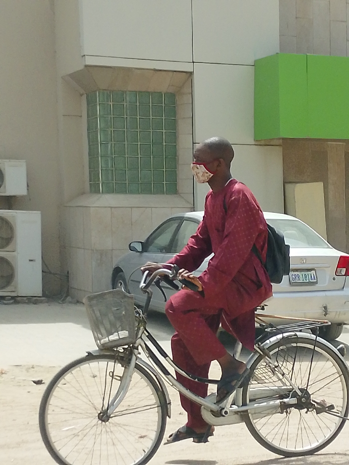 An adult male is depicted riding a silver-framed bicycle on a light-colored, unpaved surface in Fagge C, Nigeria. The individual has a shaved head and wears a maroon traditional tunic and matching trousers, dark sandals, and a patterned, light-colored fabric face mask covering his nose and mouth. A black backpack is worn over his left shoulder. The bicycle features a wire basket on the front handlebars and a rear cargo rack. In the background, a silver Honda Civic sedan, bearing Nigerian license plate GRR-181AA, is parked. To the left of the frame, a light-colored building with beige stone detailing presents a large rectangular window constructed from green glass blocks. Two white exterior air conditioning units are mounted on this building's wall. A section of a building with bright green paneling is visible on the far right. The scene is illuminated by direct sunlight.
