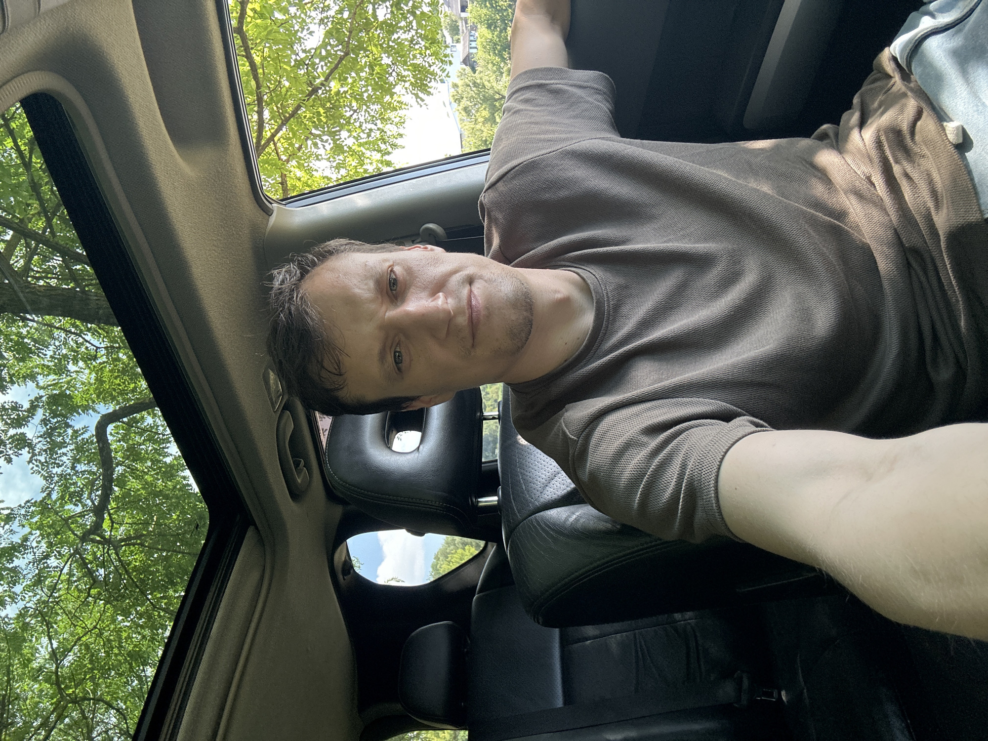 A man is taking a selfie inside a car. He is in the driver's seat, with the car's sunroof open and trees visible overhead. The background shows a residential area with houses and more trees. The man is wearing a brown t-shirt and jeans. The image was taken in Makiv, Ukraine.