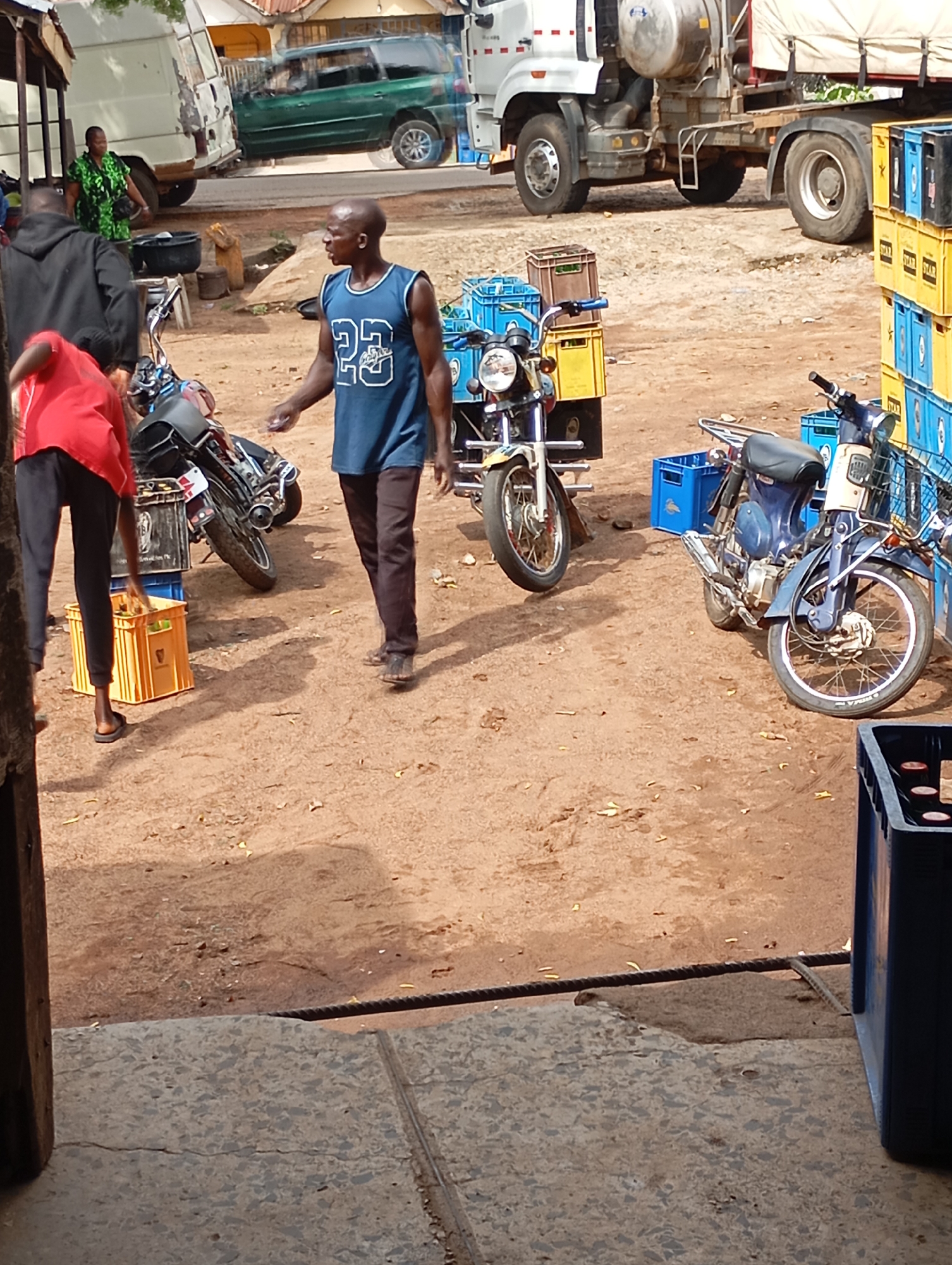 The image depicts an outdoor scene in the city of Shendam, Nigeria, likely a marketplace or a roadside area. In the foreground, a man wearing a blue sleeveless jersey with the number "23" and dark trousers stands in the center of the frame. To his left, another person in a red shirt and black trousers is bent over, interacting with yellow crates. To the right of the central figure, a motorcycle is parked, with another motorcycle partially visible in the background.
The environment is characterized by a dusty, dirt ground. In the background, various vehicles are present, including a white truck with a large tank, a green SUV, and a white van. Stacks of blue and yellow crates are visible, suggesting commercial activity. The lighting suggests it is daytime. The overall atmosphere appears to be busy and functional, with individuals engaged in tasks related to the transport and storage of goods.