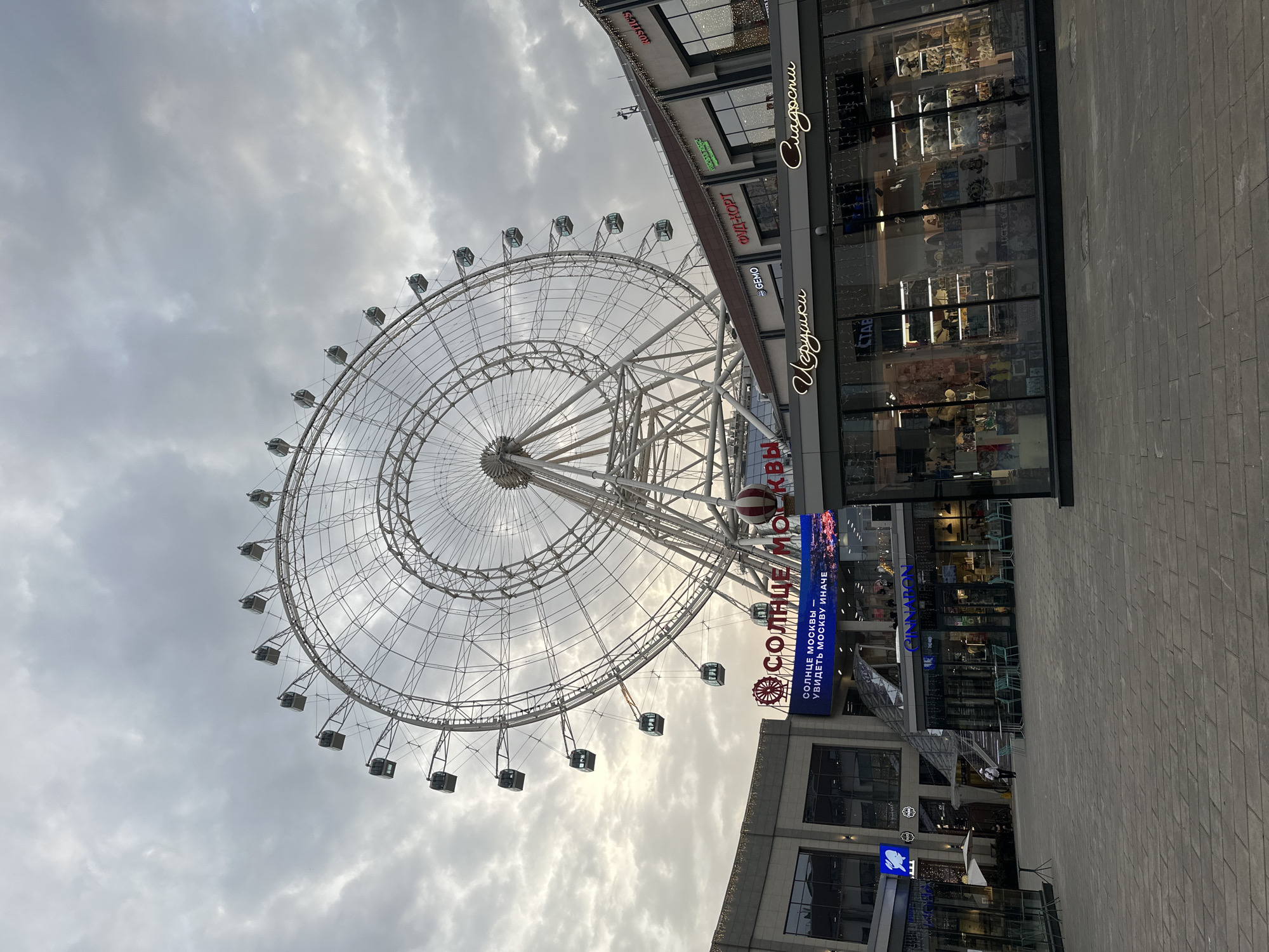The image depicts a large Ferris wheel against a cloudy sky, located next to a commercial building in Dolgoprudny, Russia. The Ferris wheel, identified as "Sun of Moscow" ("Солнце Москвы" in Russian), is white with enclosed cabins. The adjacent building features signage in Russian, with shops including "Sweets" ("Сладости"), "Toys" ("Игрушки"), "Food Court" ("ФУДІ-КОРТ"), and "CINNARON." The ground is paved with gray bricks. There are no people visible in the immediate foreground, but some indistinct figures may be present near the building entrance.