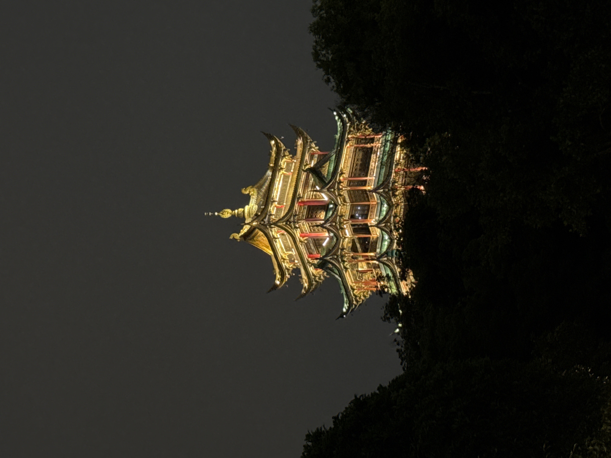 The image shows a brightly illuminated, multi-tiered pagoda at night, located in the city of Liangjiang, China. The building features intricate golden details and is situated amid dark foliage, likely trees. The sky appears overcast or dark. The composition suggests the pagoda is the main subject, illuminated against a backdrop of darkness.