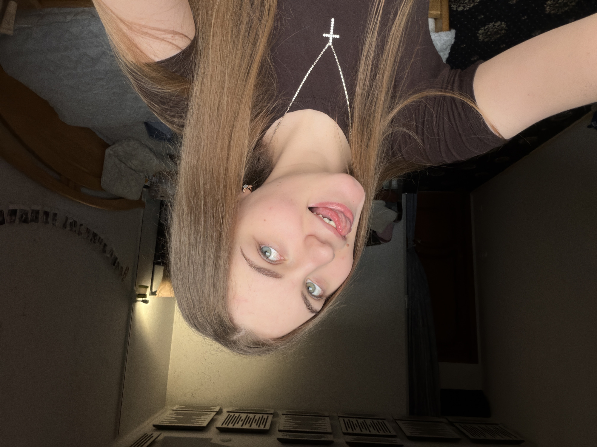 A young woman with long, blonde hair and blue eyes is taking a selfie in a dimly lit room. She is wearing a brown t-shirt and a silver necklace with a cross pendant. Her tongue is sticking out playfully. The background shows a bed with a gray comforter, a wooden headboard, and a wall decorated with string lights and small photos. In the distance, a door and a closet are visible. The ceiling has square acoustic panels. The scene appears to be a bedroom, likely in a residential apartment or house, located in Dnipro, Ukraine, as suggested by the context provided.