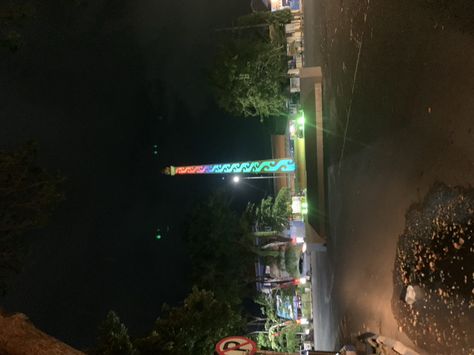 The image shows a tall, thin, brightly colored column illuminated by a string of LED lights, in the middle of a small, dark, and empty square in Probolinggo, Indonesia. The column has a distinct wave pattern, painted in shades of red, pink, green, and turquoise. The square is bordered by a low, flat, pale-colored wall, with a small, paved road on the left side. The road is dark, and the only light is from the column and a streetlight in the background. Several trees are visible, their leaves are dark and indistinguishable. Some cars are parked in the background, and a "no parking" sign is seen on a tree on the left.
