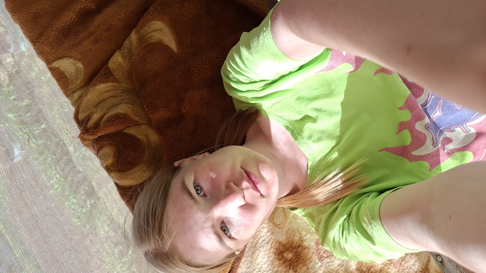 A woman lies supine on a bed covered with a brown patterned blanket. She wears a light green long-sleeved shirt with a pink and blue design. Her blond hair is partially visible. The photo appears to be taken at an oblique angle, capturing the upper portion of her body. The environment includes a translucent curtain or window covering, partially visible in the upper left corner. No specific identifying features indicate the precise location in Большой Букор, Russia.